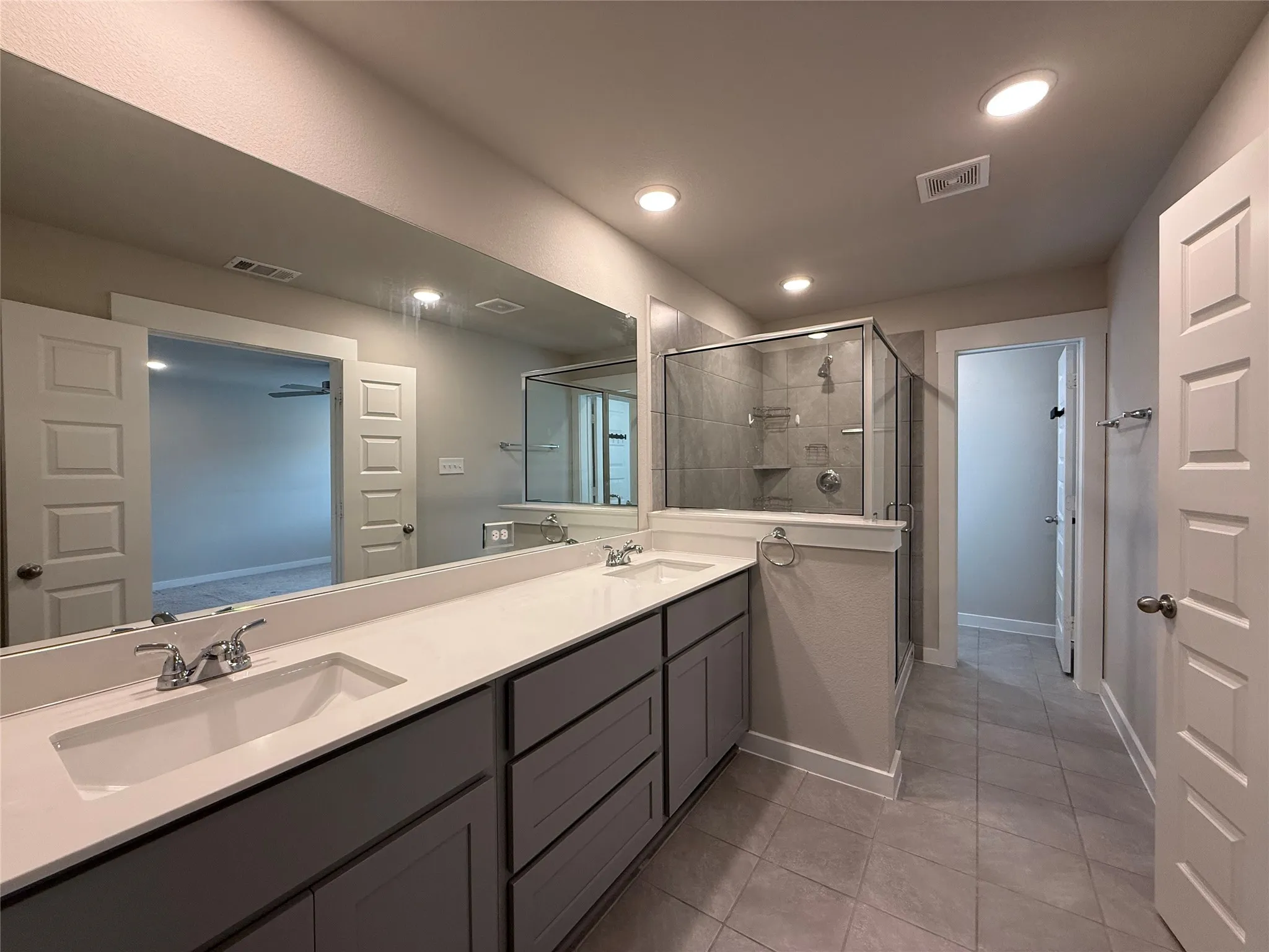 Dual vanities with undermount sinks and chrome fixtures, quartz countertops, gray cabinetry, tile flooring, and a glass-enclosed shower with tile surround