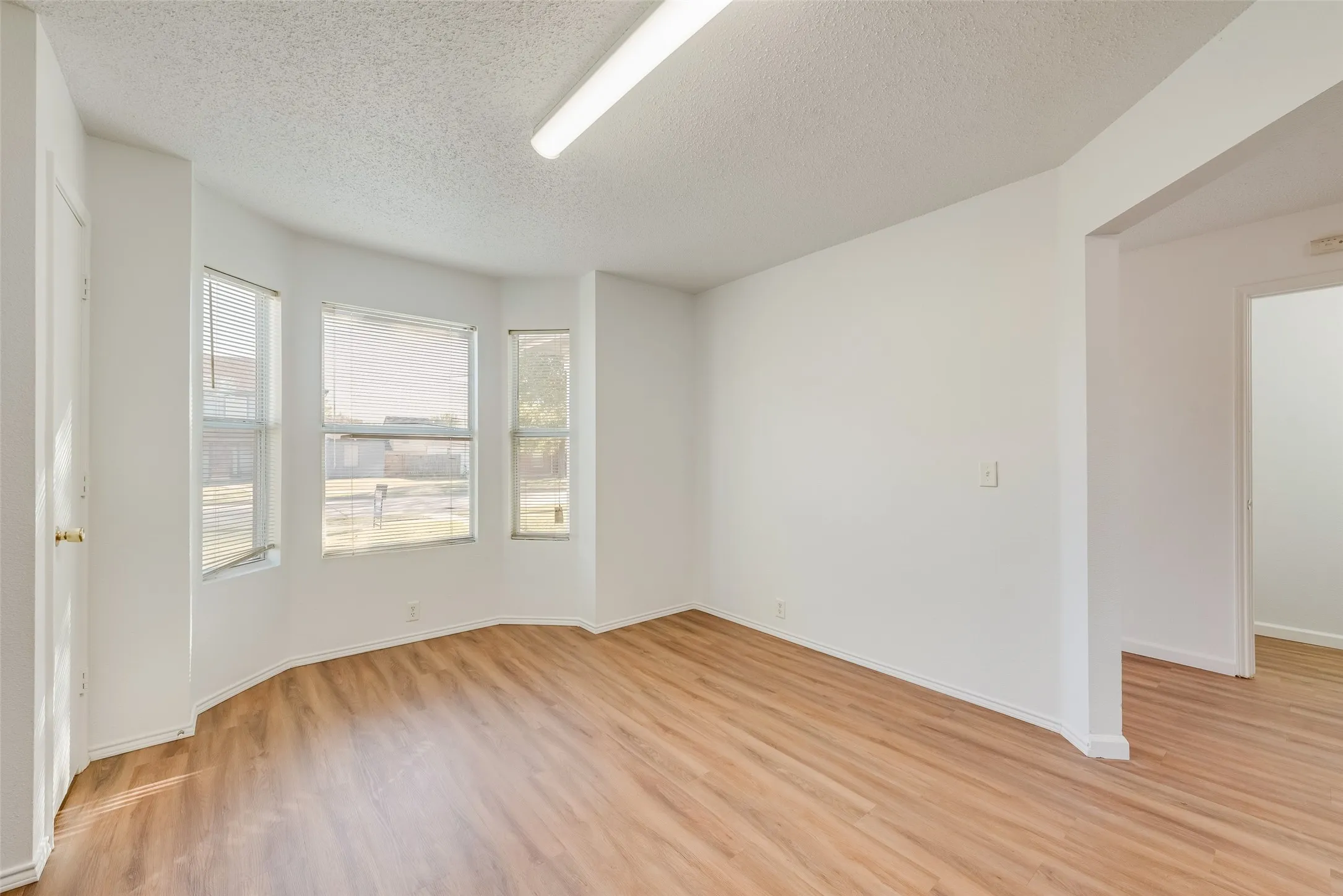 Bay window with three windows, wood-finish flooring, white walls, and a fluorescent ceiling light fixture