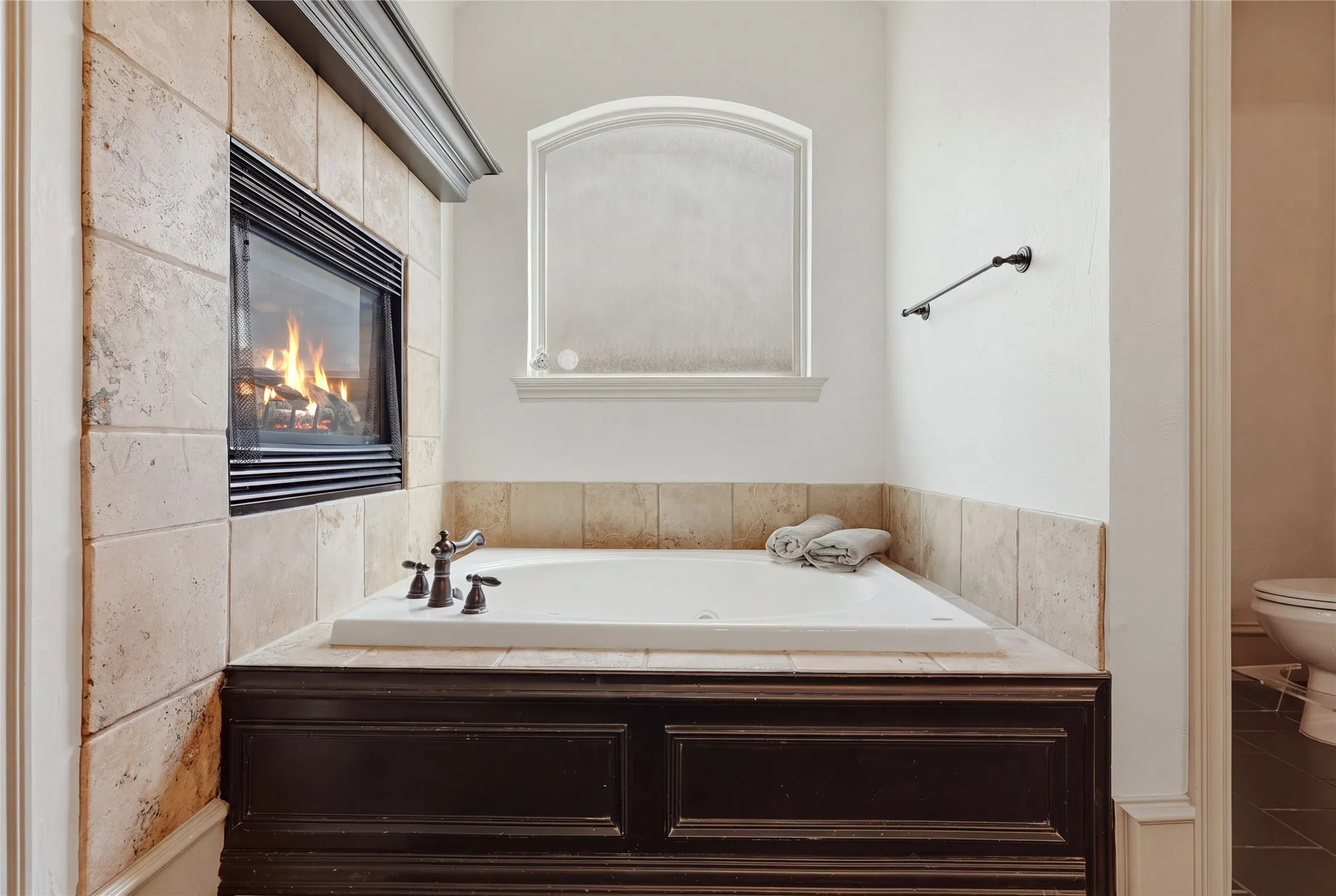 Built-in soaking tub with dark wood-finish paneling, an integrated fireplace with tile surround, and an arched privacy window