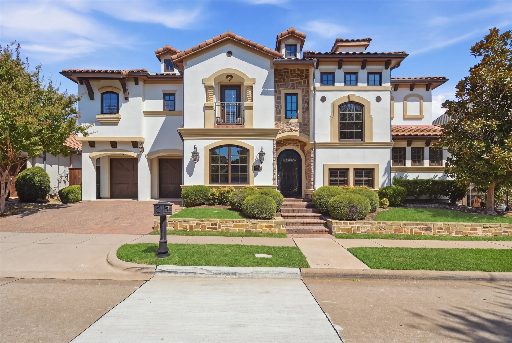 Stucco exterior with stone accents, arched windows, tile roof, dual garage doors, and a stone retaining wall