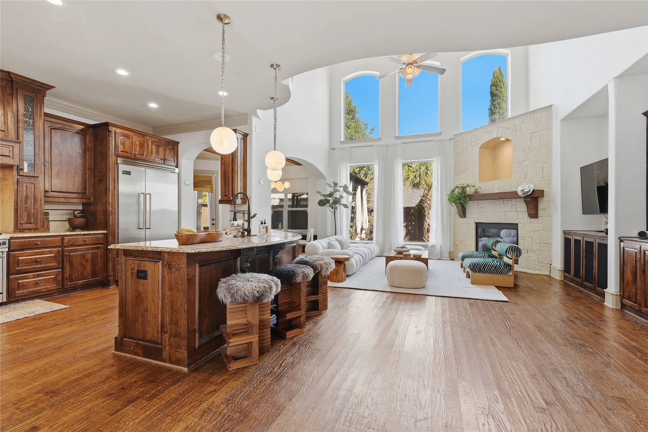 Open-concept living space with wood-finish flooring, a stone-clad fireplace with mantel, and a kitchen island featuring pendant lighting