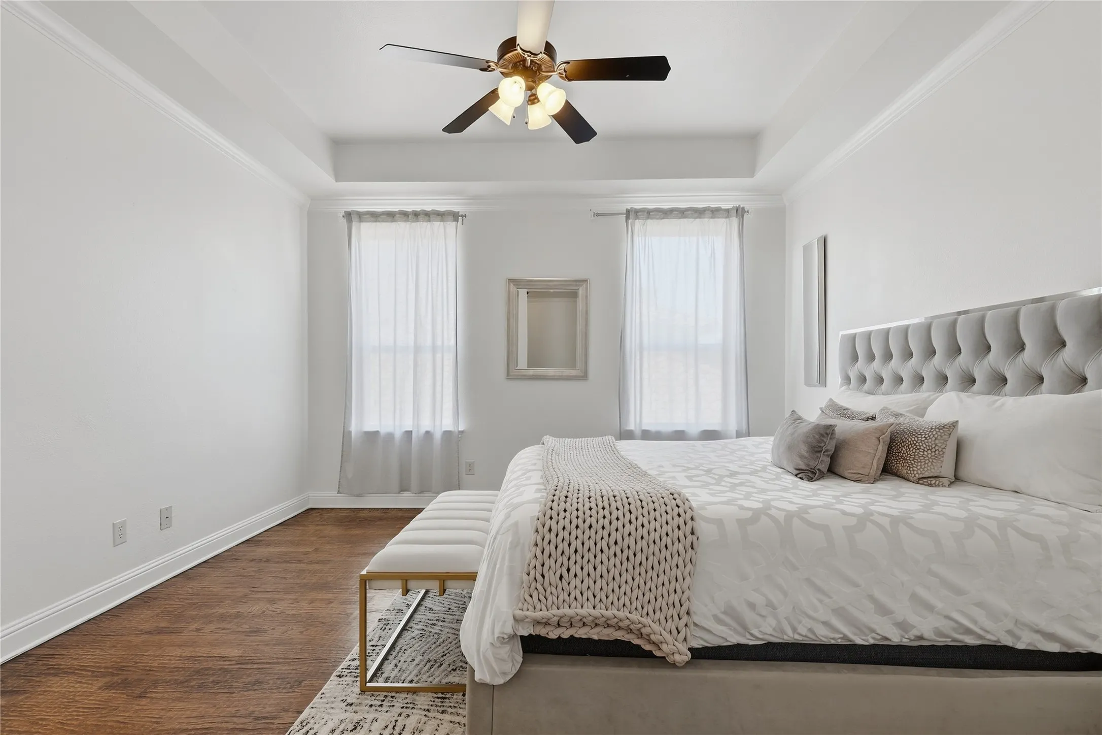 Tray ceiling with a ceiling fan, two windows with sheers, wood-finish flooring, and crown molding