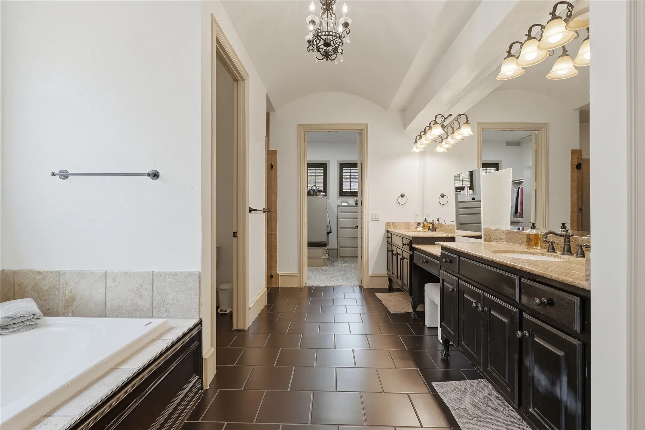 Dual vanities with stone countertops, dark wood cabinetry, a built-in tub with tile surround, and vaulted ceilings with a chandelier