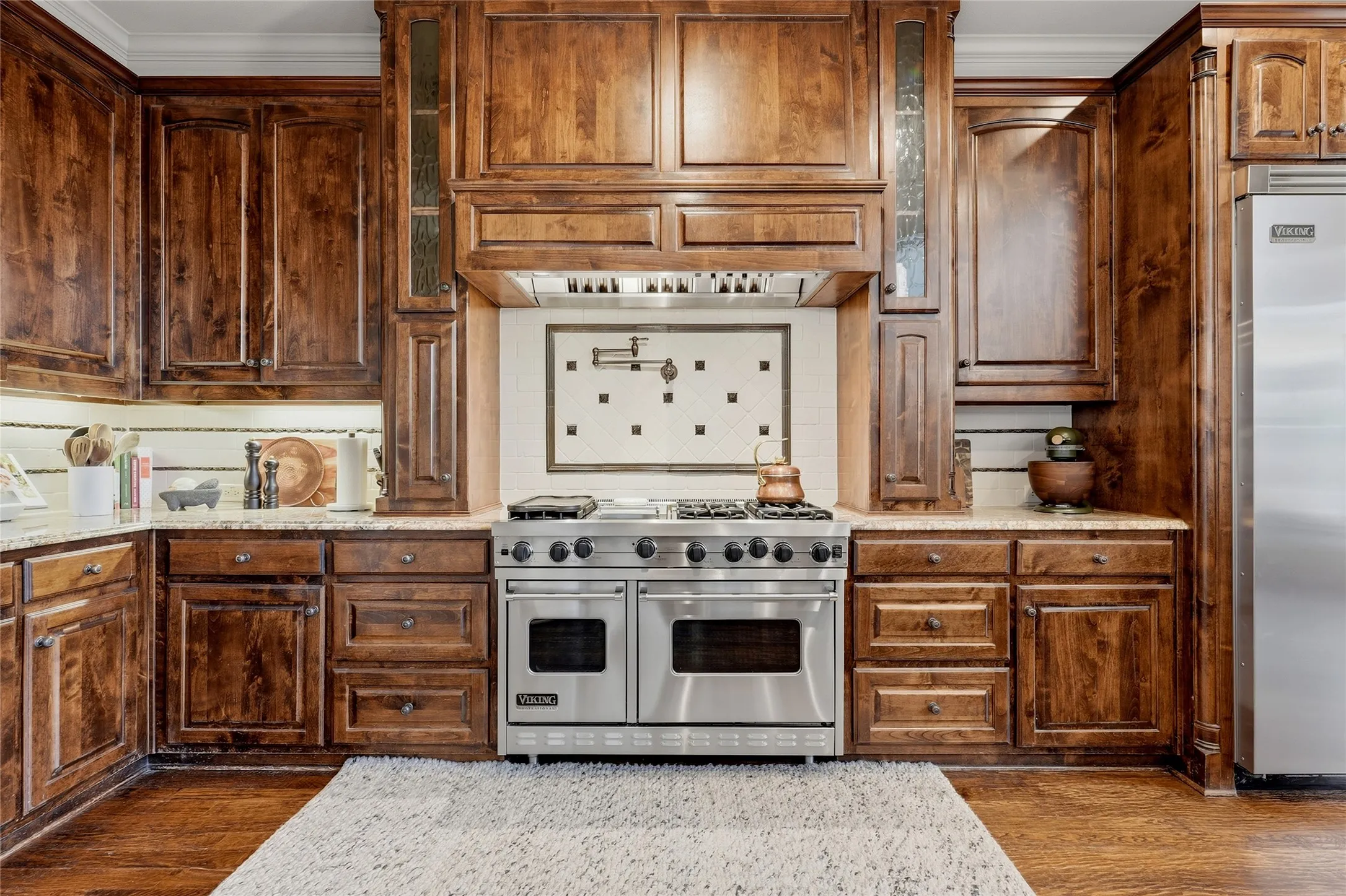 Rich wood cabinetry with varied door styles, stainless steel professional-grade range, tiled backsplash with decorative inset, under-cabinet lighting, and light-colored countertops