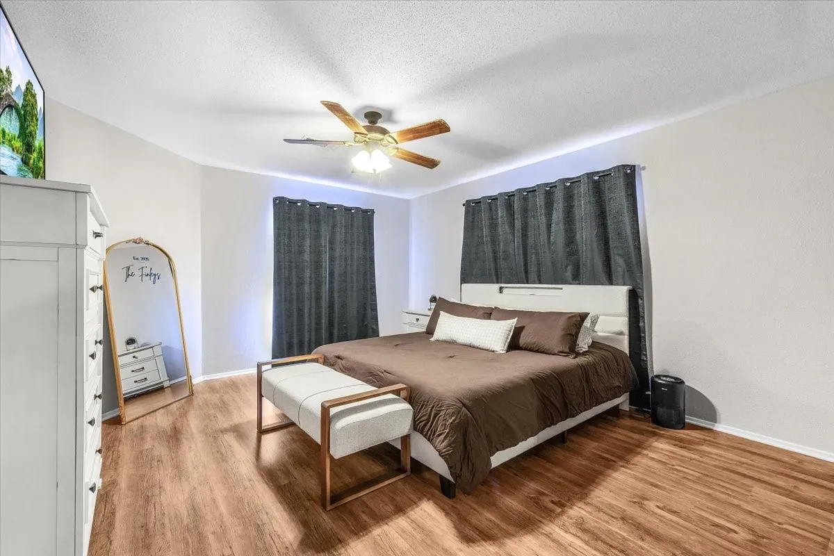 Spacious primary bedroom wood-finish flooring throughout, bright white walls, and a ceiling fan with integrated lighting