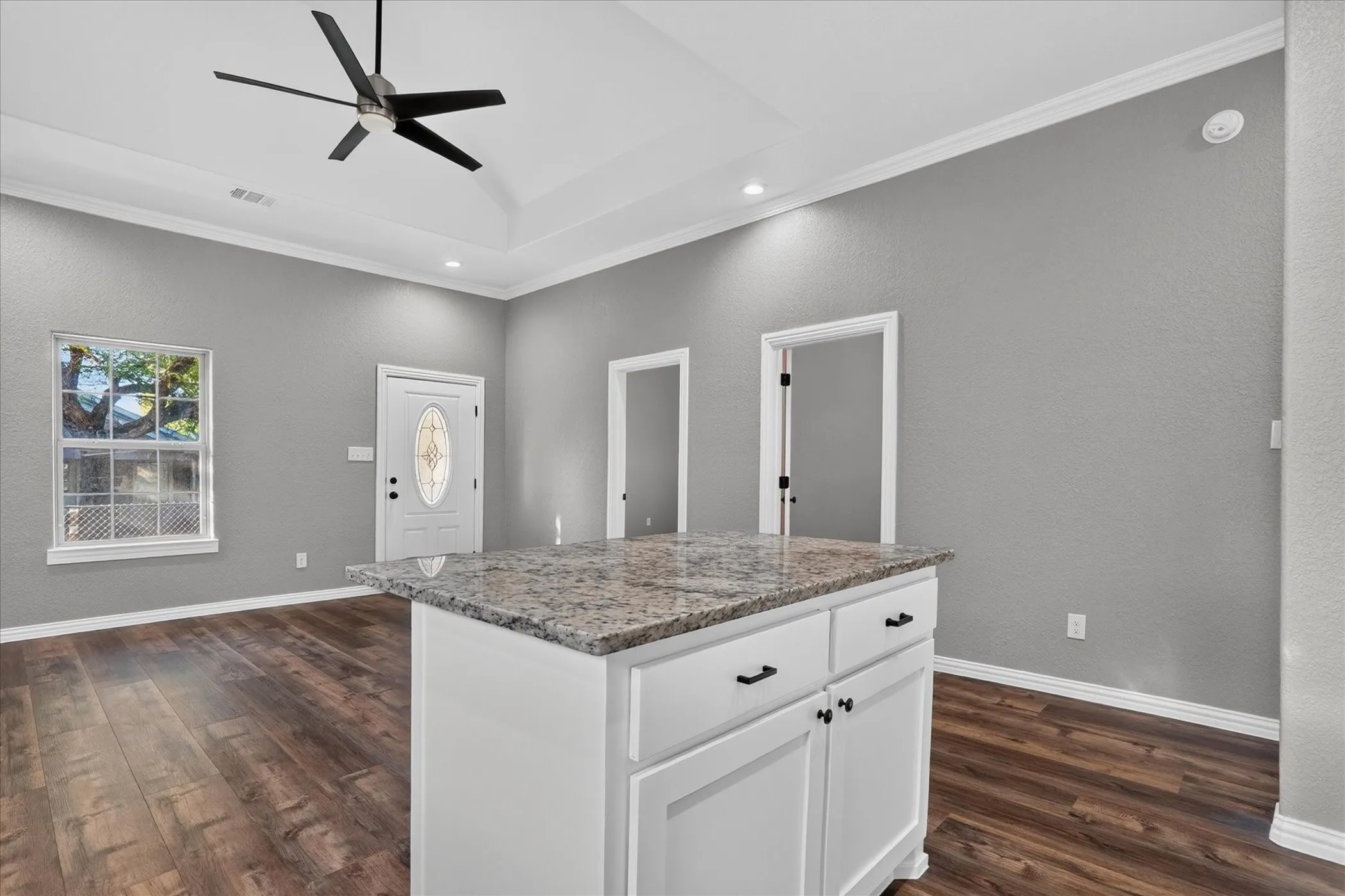 Kitchen island with stone countertop and white cabinetry, tray ceiling with recessed lighting and ceiling fan, wood-finish flooring, and a decorative glass front door