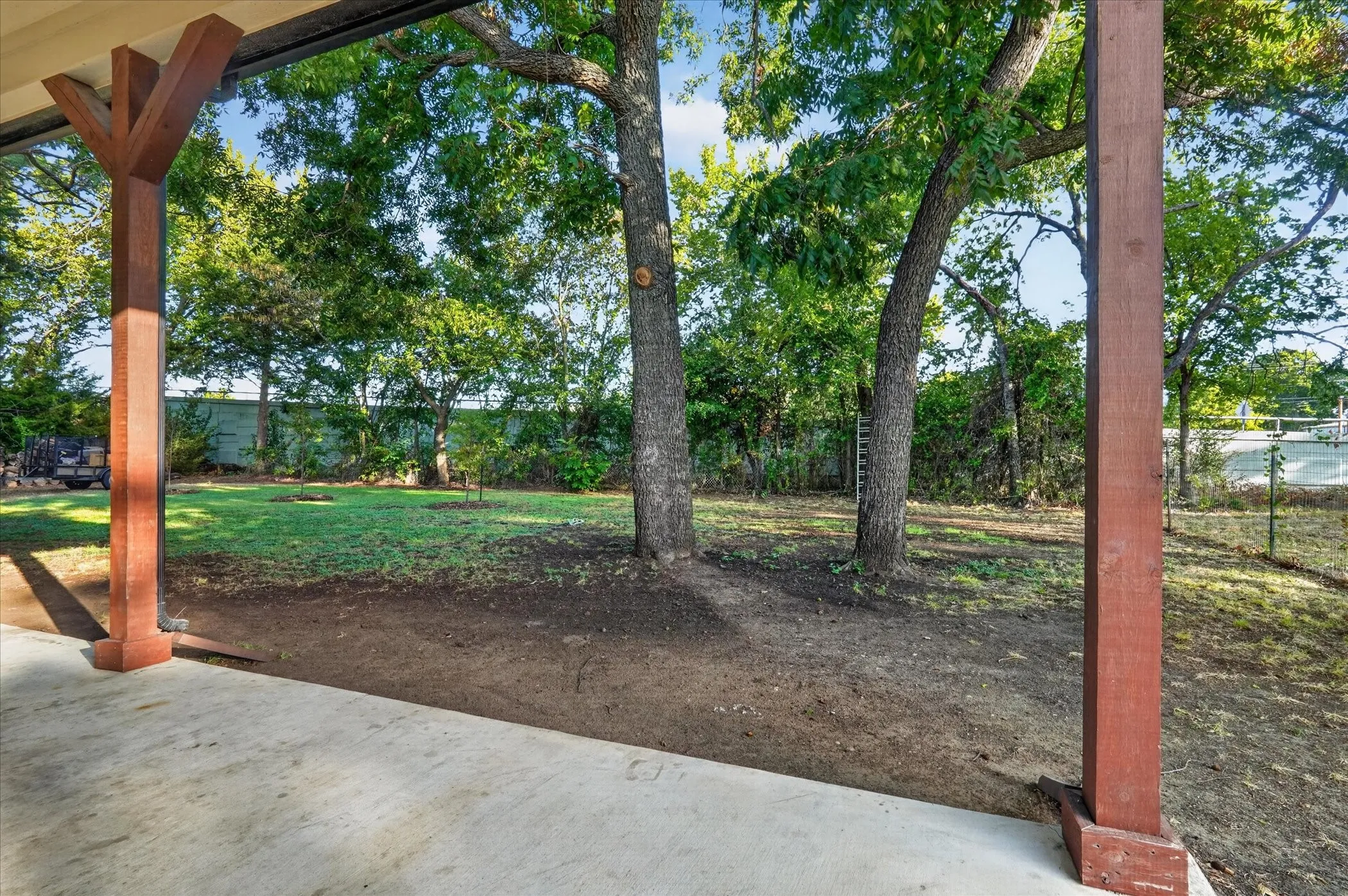 Covered patio with concrete flooring, supported by substantial wood columns, overlooking a grassy yard with mature trees and perimeter fencing