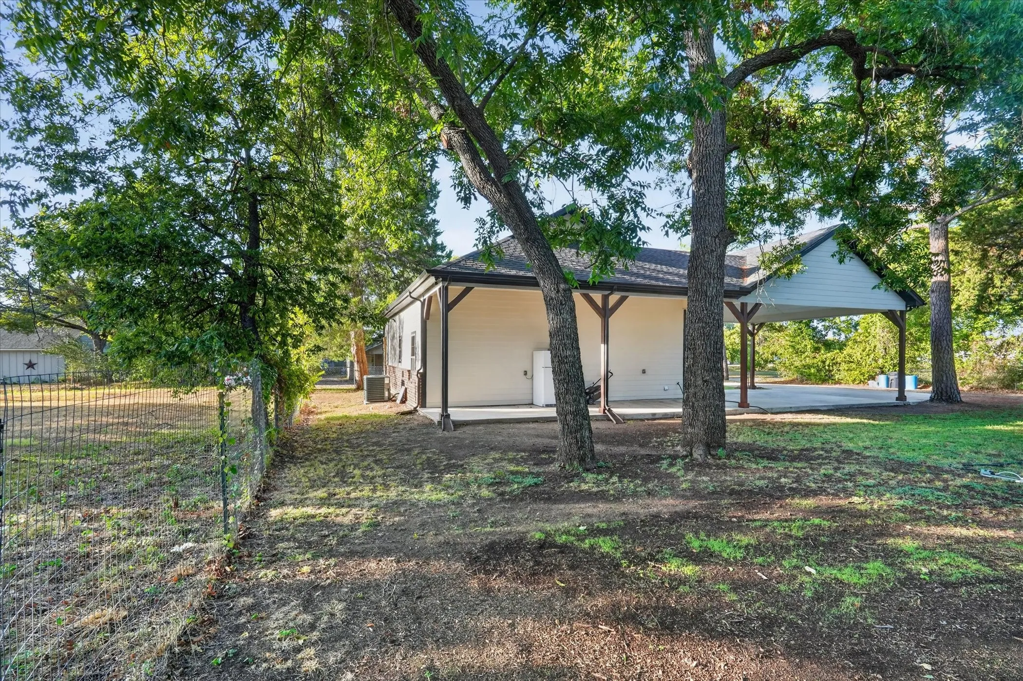 Attached carport with gabled roof, covered patio space, and mature trees