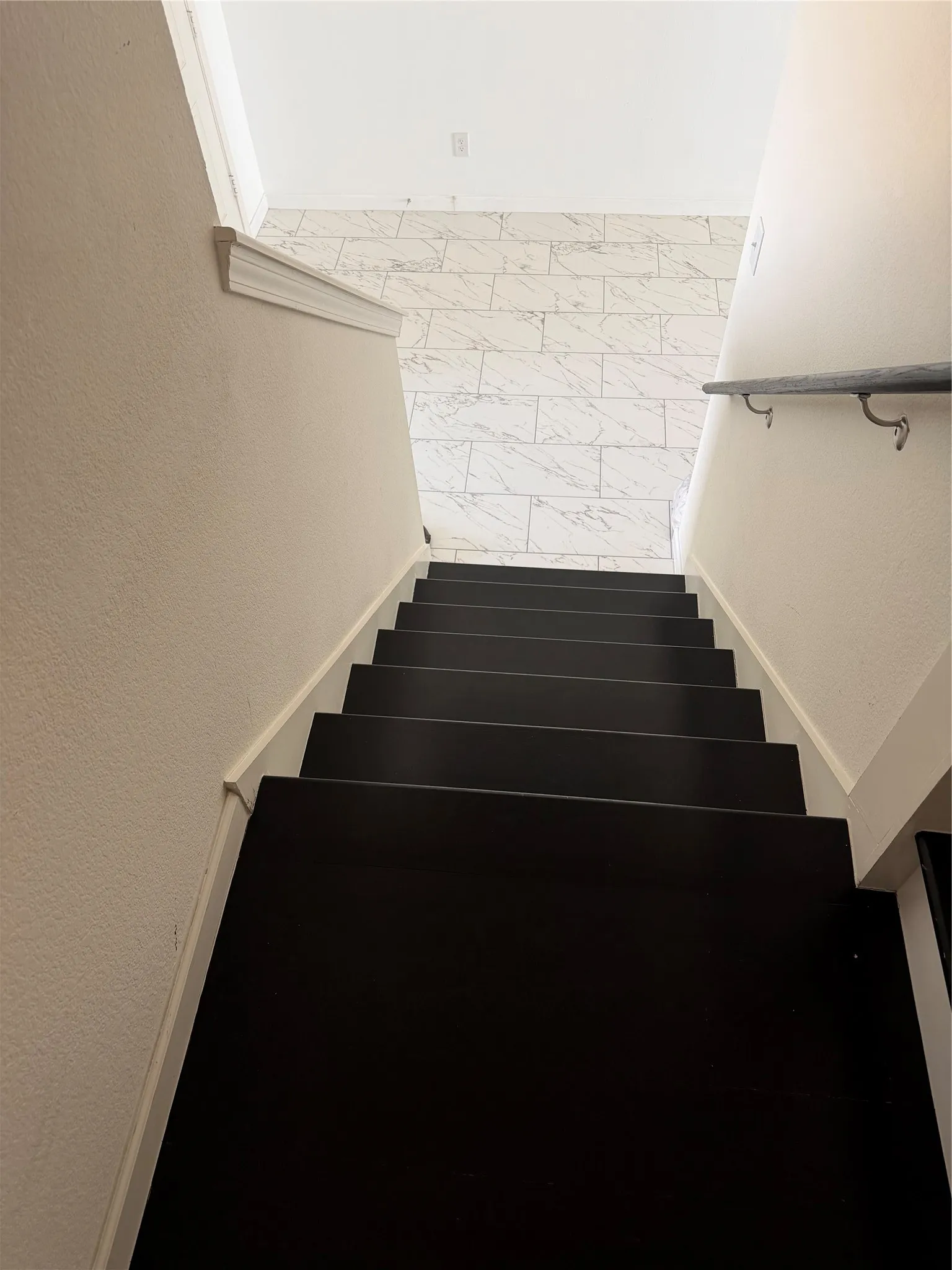 Dark-finished staircase with a wood handrail, leading to a space with tile flooring