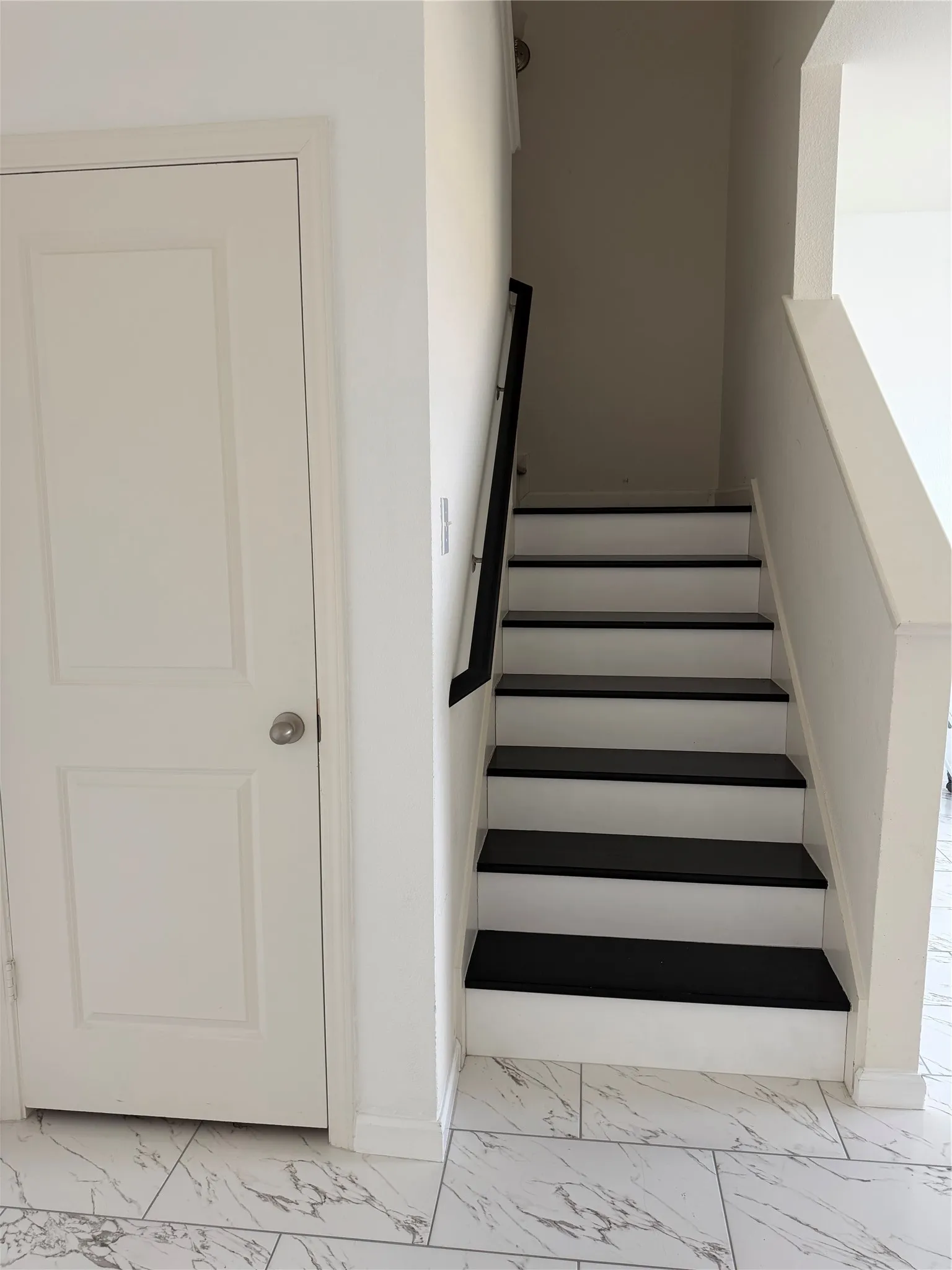 White and black staircase with a dark handrail, white interior door, and marble-look tile flooring