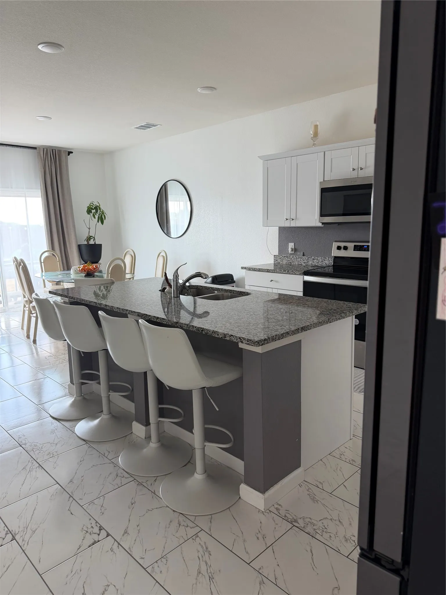 Kitchen island with a granite countertop and integrated sink, white cabinetry with stainless steel appliances, and large format tile flooring