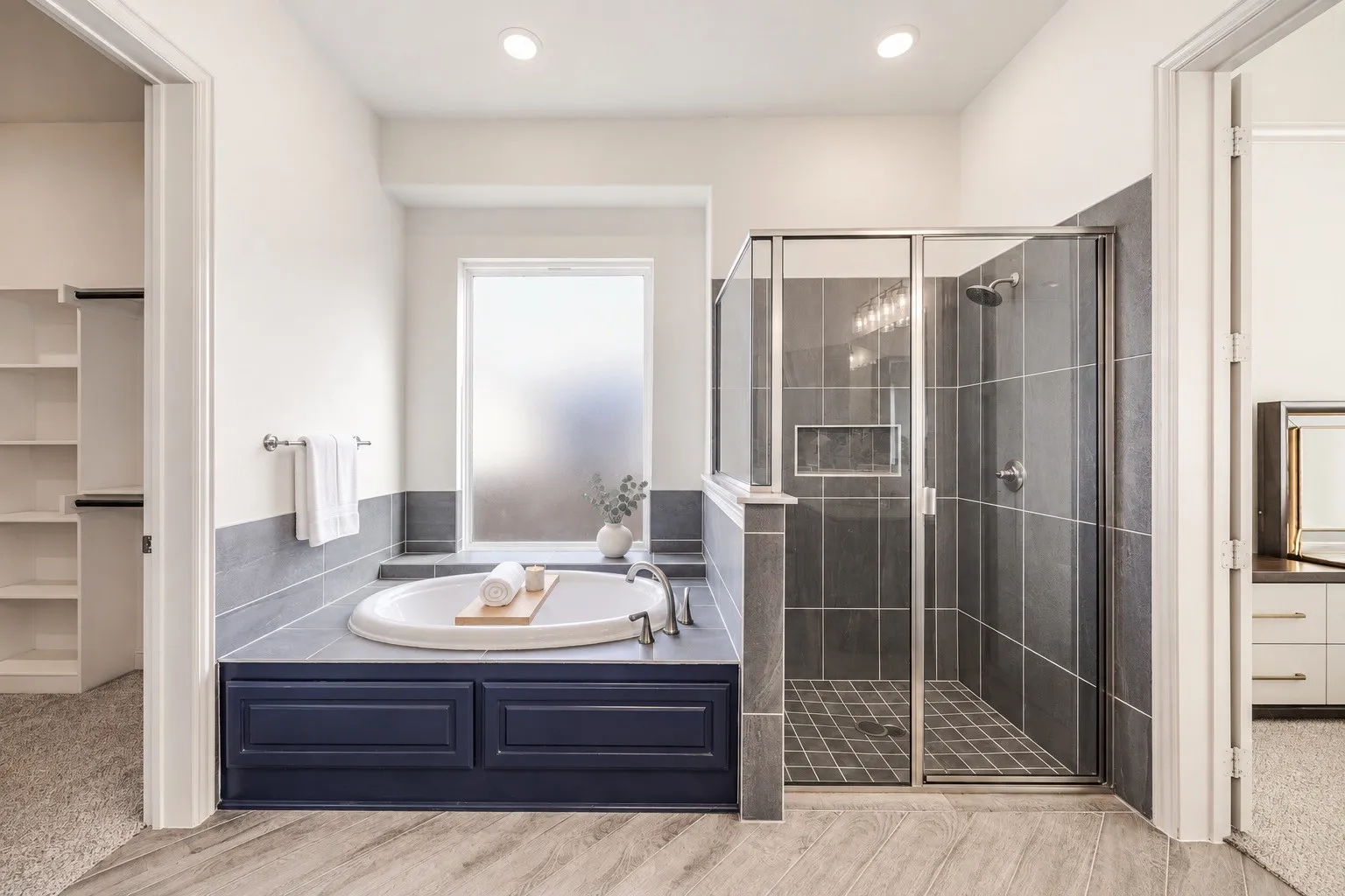 Recessed tub with dark blue paneling, glass-enclosed shower with tile surround, wood-finish flooring, and a walk-in closet with built-in shelving -staging with AI