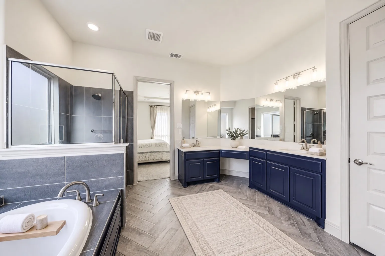 Spacious bathroom featuring a freestanding soaking tub, separate glass-enclosed shower with gray tile surround, dual vanity with blue cabinetry, white countertops, and herringbone-patterned floor tile -staging with AI