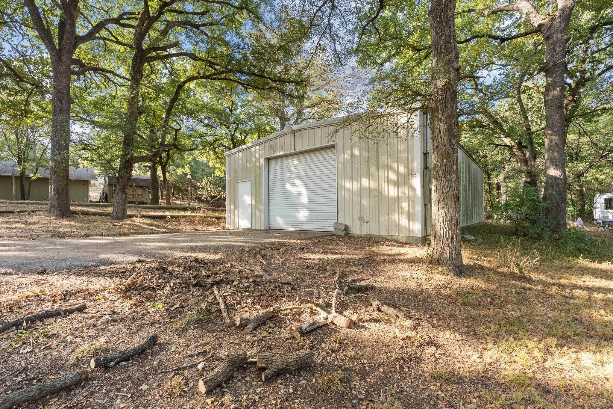Metal-sided outbuilding with a roll-up garage door and an entry door, surrounded by mature trees