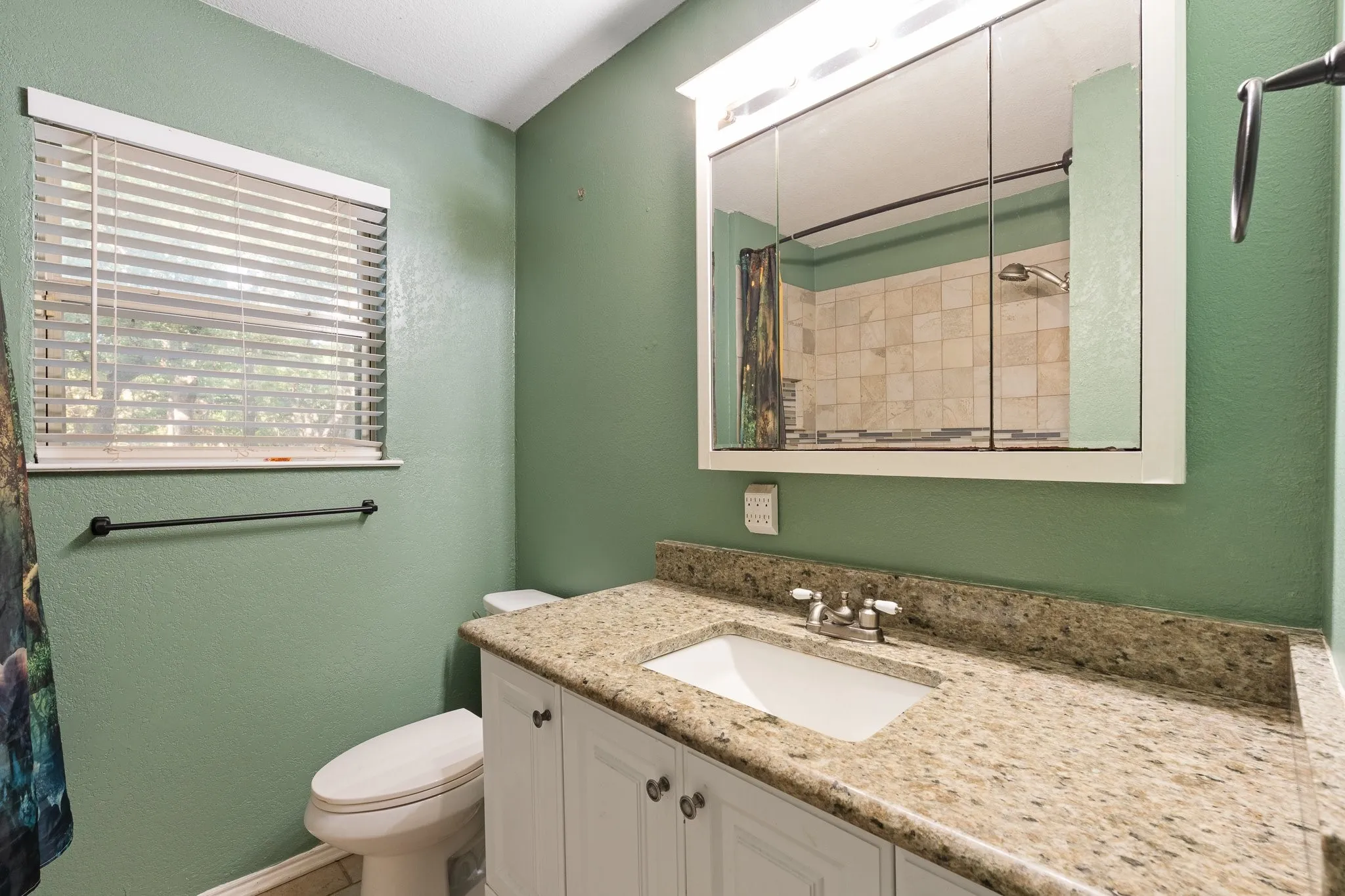 Vanity with stone countertop and integrated sink, white cabinetry with dark hardware, mirrored medicine cabinet with overhead lighting, and window with blinds