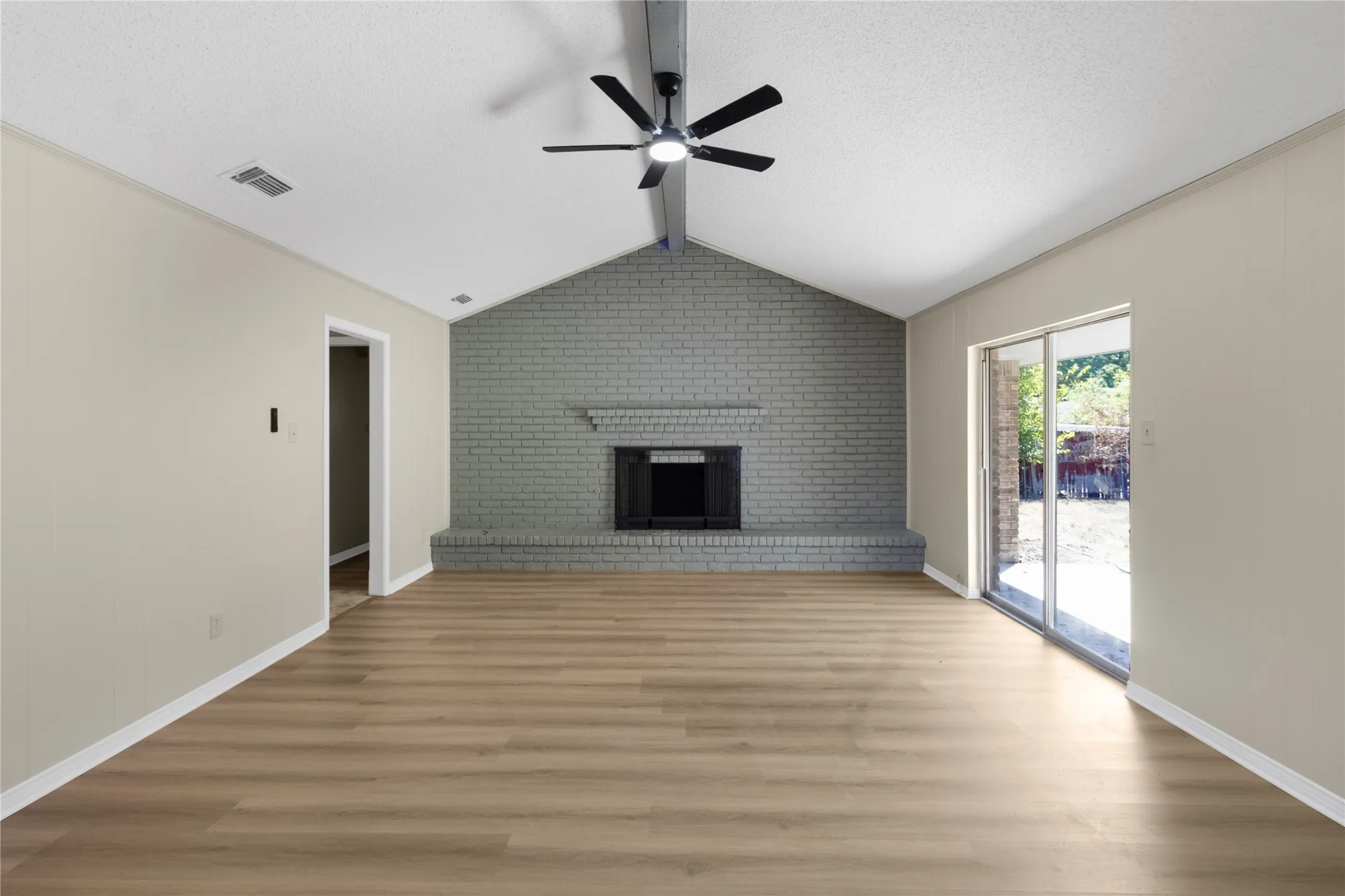 Vaulted ceiling with exposed beam, brick fireplace with mantel, wood-finish flooring, sliding glass door, and ceiling fan with integrated lighting