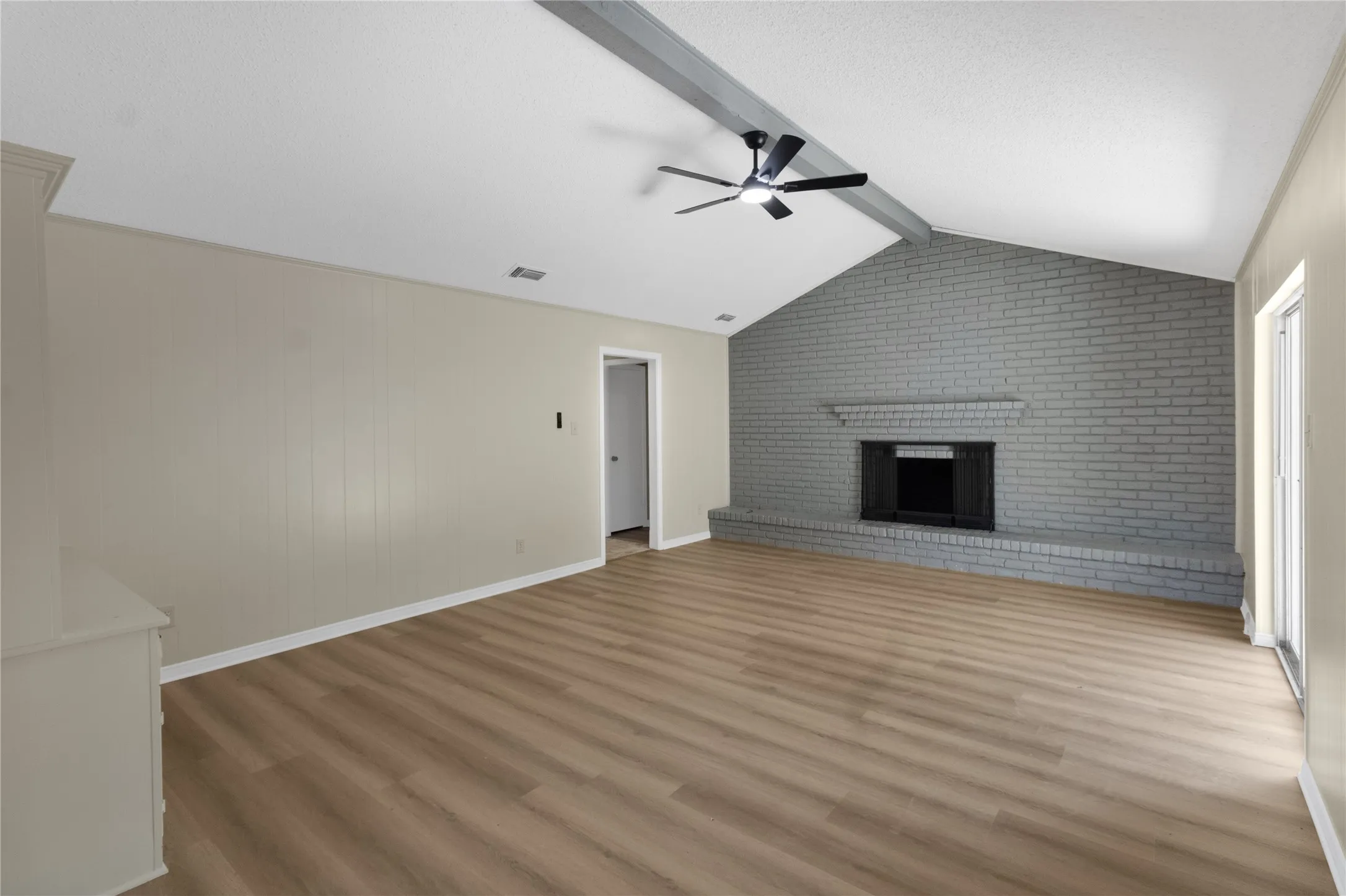 Vaulted ceiling with exposed beam, wood-finish flooring, brick fireplace with hearth, and ceiling fan