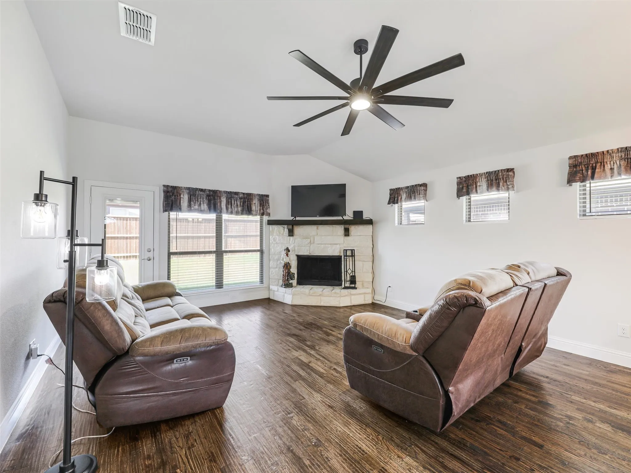 Vaulted ceiling, wood-finish flooring, corner stone-surround fireplace, large ceiling fan, and multiple windows