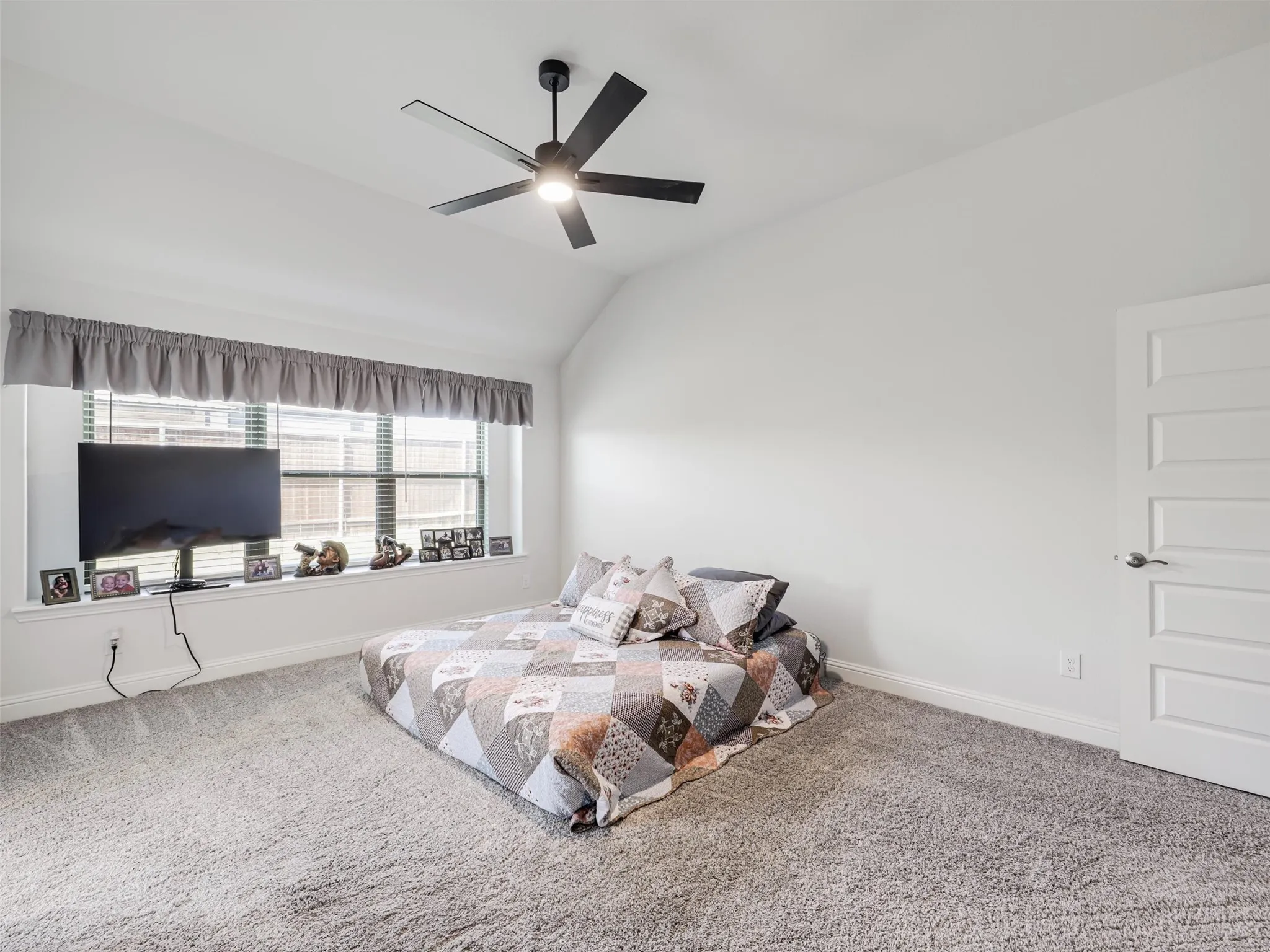 Vaulted ceiling featuring a ceiling fan with integrated lighting, large window with blinds, plush carpet flooring, and a white paneled door