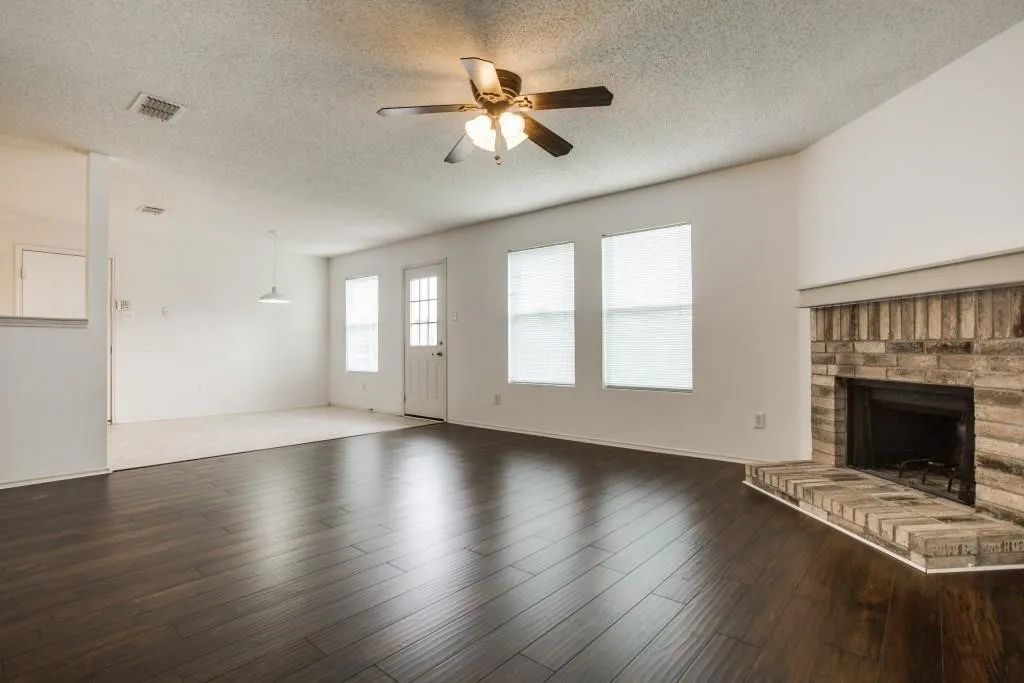 Dark wood-finish flooring, brick fireplace, multiple windows with blinds, ceiling fan with integrated lighting, and a white paneled door with glass inserts