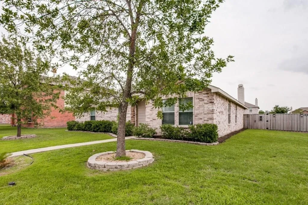 Brick exterior, mature trees with stone-lined bases, green lawn, established landscaping, and a privacy fence