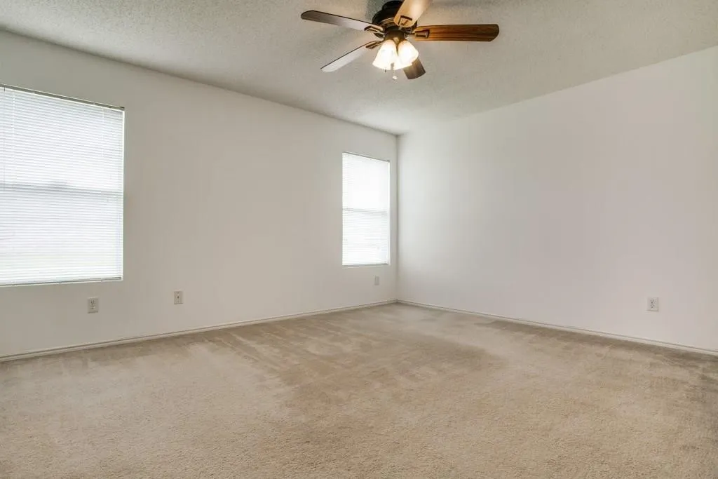 Spacious room featuring neutral carpeting, white walls, two windows with blinds, and a ceiling fan with integrated lighting