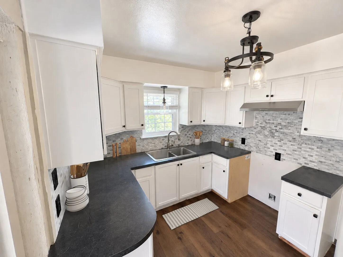 Virtually staged -
White cabinetry, dark countertops, wood-finish flooring, a mosaic tile backsplash, and a dual-basin sink