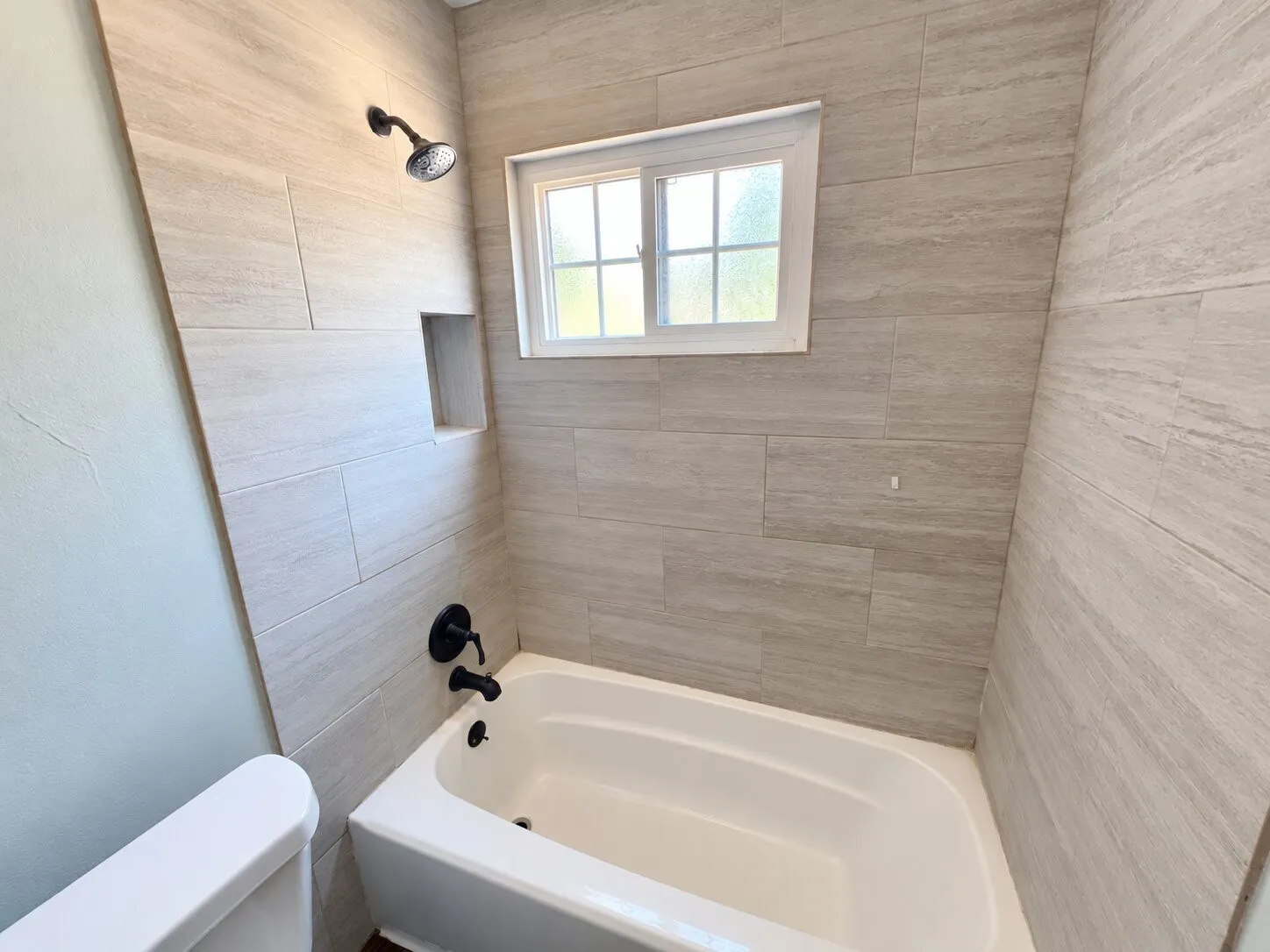 Bathtub with matte black fixtures, an integrated shower niche, and rectangular window, surrounded by light-toned tile