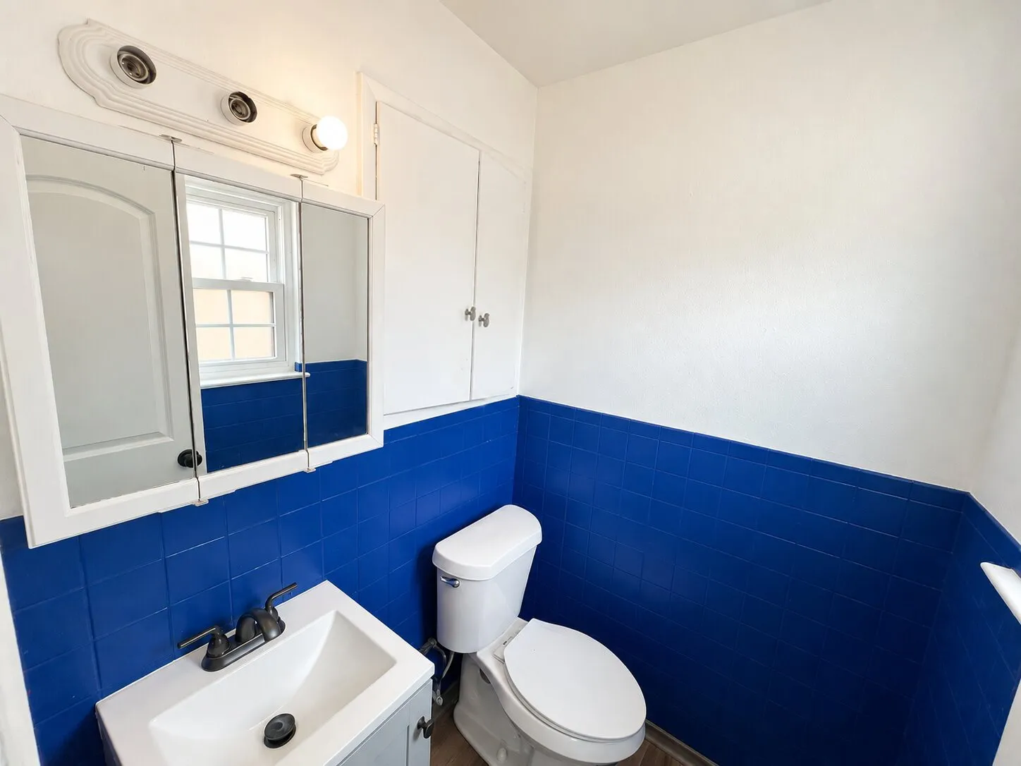Blue wall tile with white upper walls, a white vanity with an integrated sink, a dark bronze-finish faucet, and a wall-mounted medicine cabinet with a light fixture