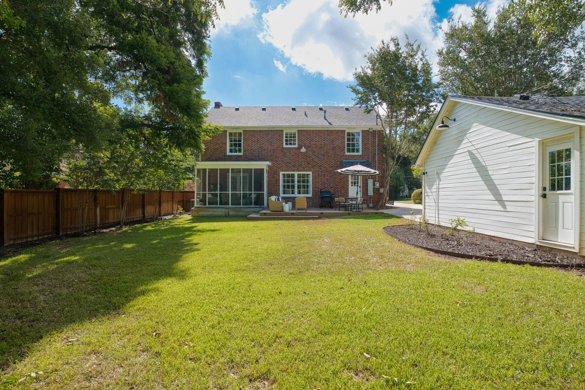 Expansive green lawn with a wood privacy fence, brick exterior, screened-in porch, and a separate white-sided auxiliary building with a door and exterior lighting