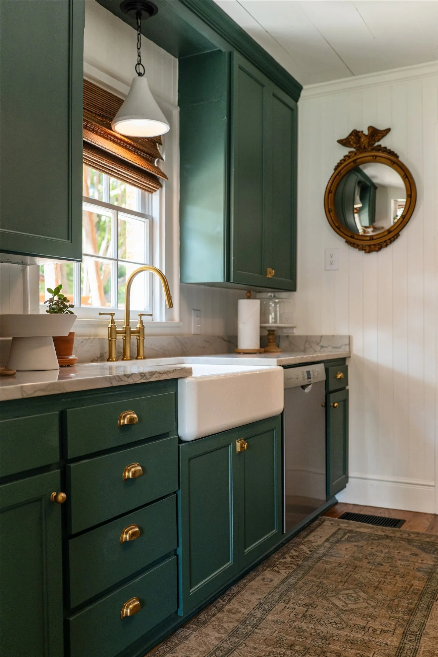 White farmhouse sink with brass hardware, dark green cabinetry, light-toned countertop, and stainless steel dishwasher