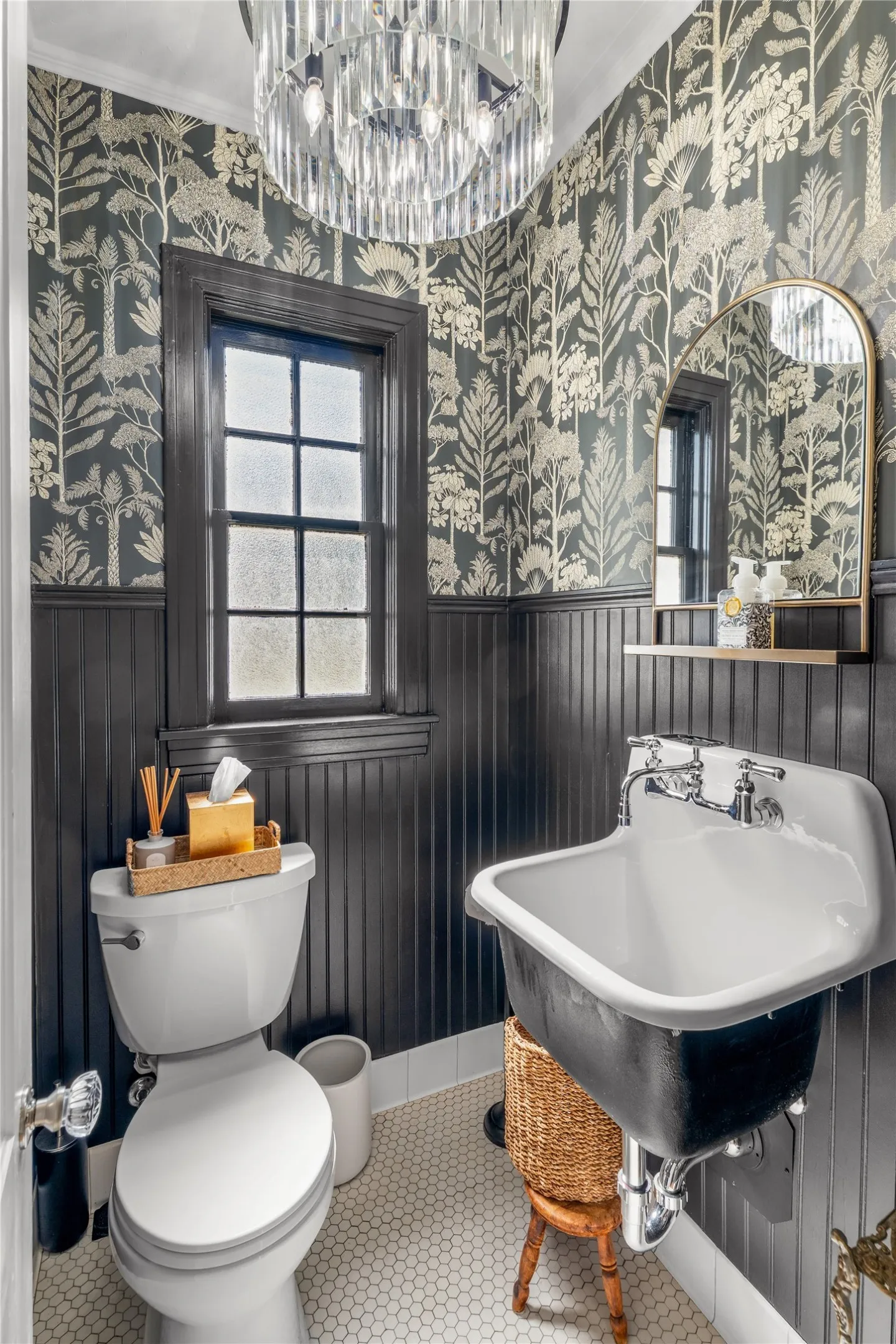Patterned wall covering, beadboard paneling, clear glass chandelier, white hexagonal floor tile, and a wall-mounted basin sink with chrome fixtures