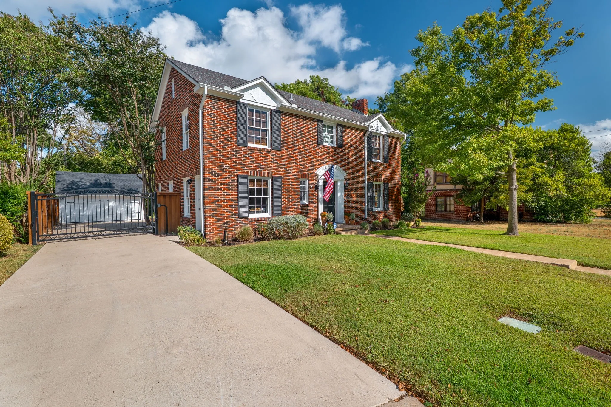 Brick exterior with white trim, a concrete driveway, a wrought iron gate, a detached structure, and a manicured lawn