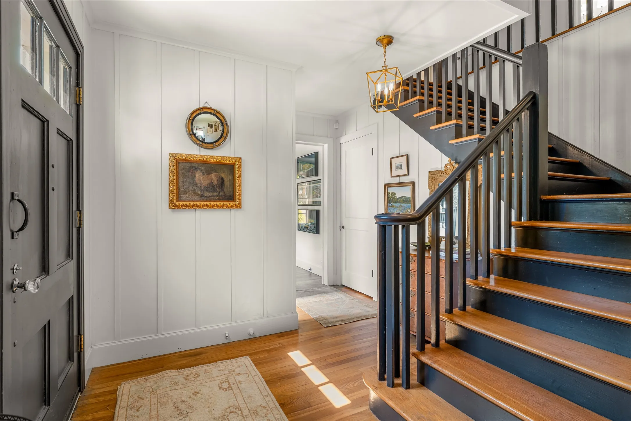 Entryway featuring wood-finish flooring, vertical paneling, a dark-toned paneled entry door with transom lites, and a staircase with wood treads and dark balusters