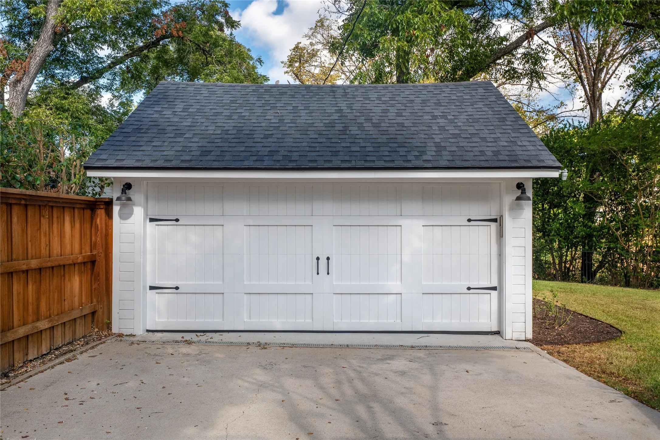 Detached garage featuring a dark shingle roof, white panel doors with decorative hardware, and integrated exterior lighting