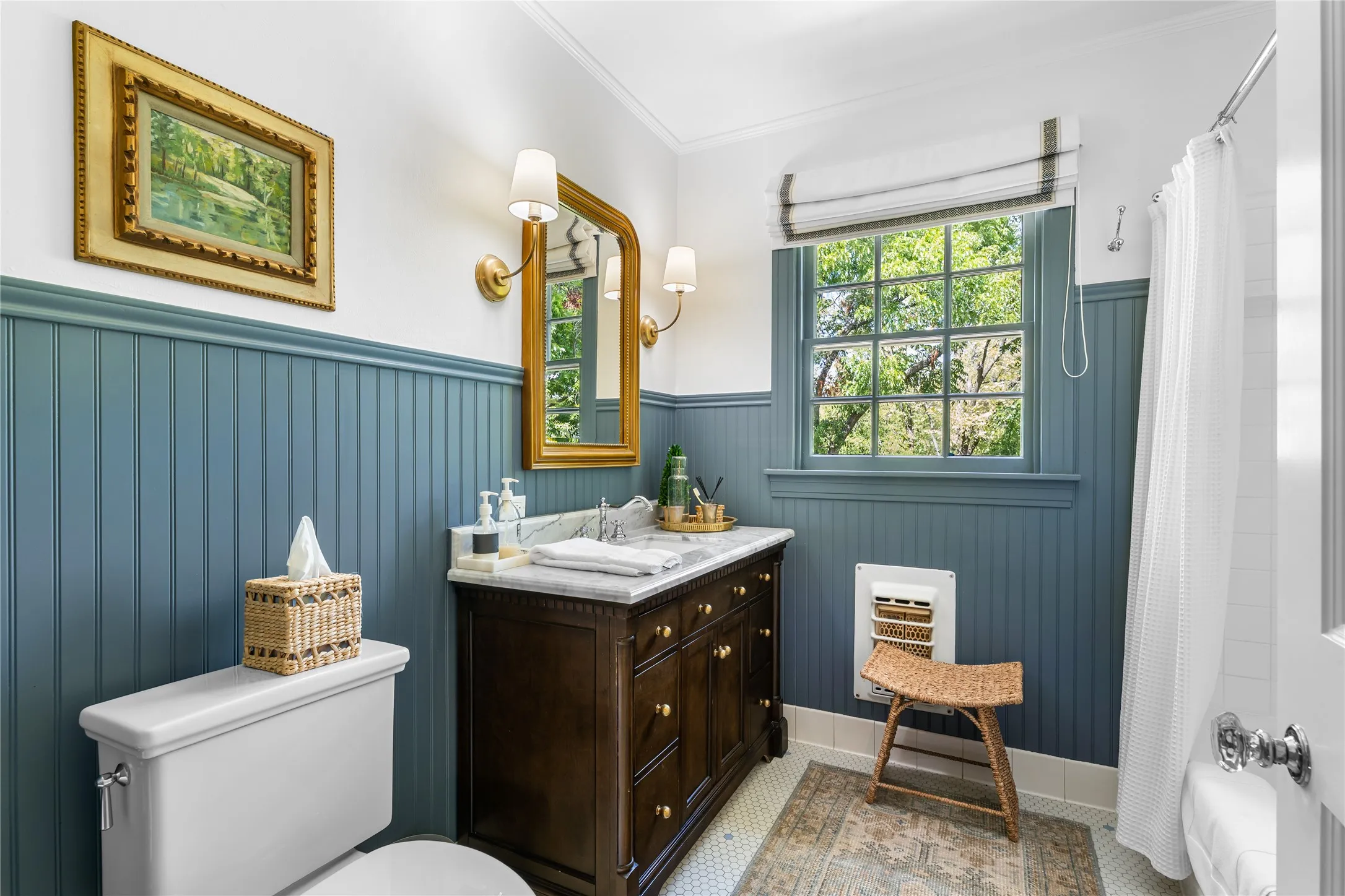 Bathroom featuring beadboard wainscoting, a dark wood-finish vanity with a stone countertop, a gold-framed mirror, and a window with a Roman shade