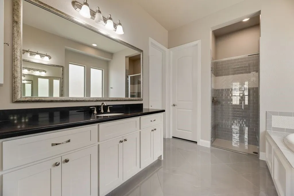 White cabinetry with dark countertop, large vanity mirror with decorative frame, glass-enclosed shower with subway tile, white soaking tub with tiled surround, and light-toned floor tile