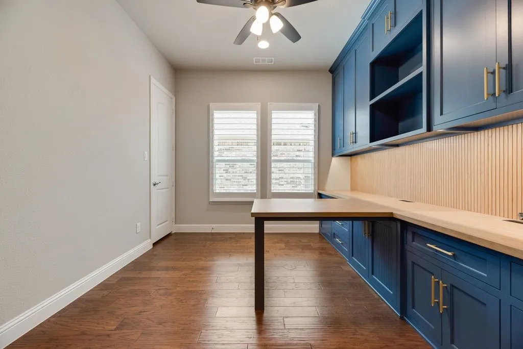 Built-in blue cabinetry with gold-tone hardware, wood-finish flooring, two windows with louvered blinds, and a ceiling fan with integrated lighting