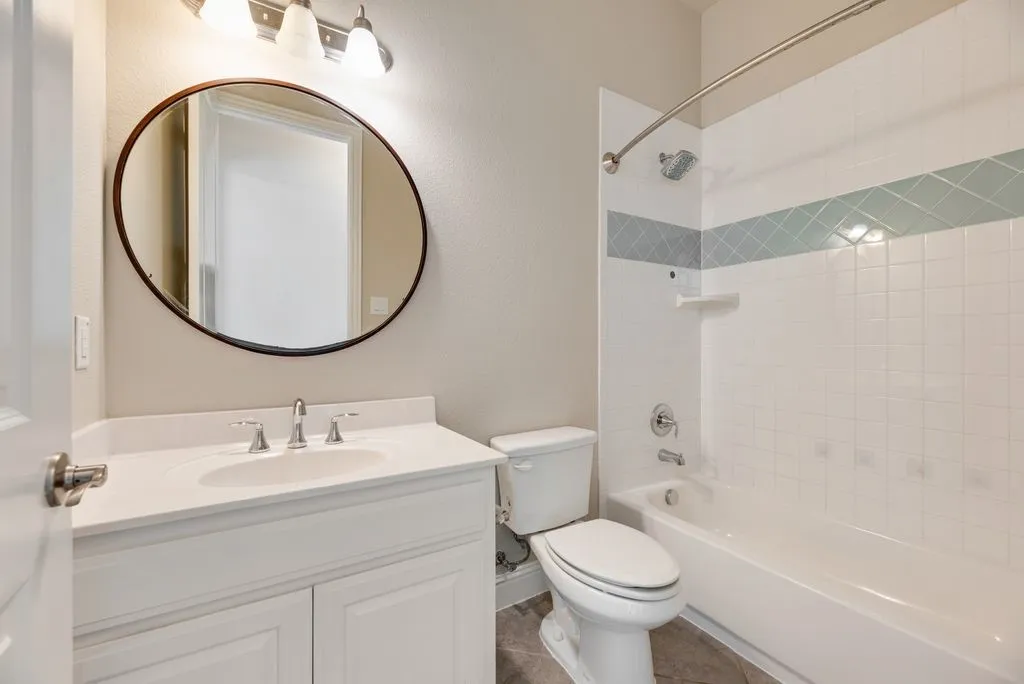 White vanity with an integrated sink and brushed nickel faucet, round framed mirror, three-light vanity fixture, bathtub with tile surround, and patterned accent tile