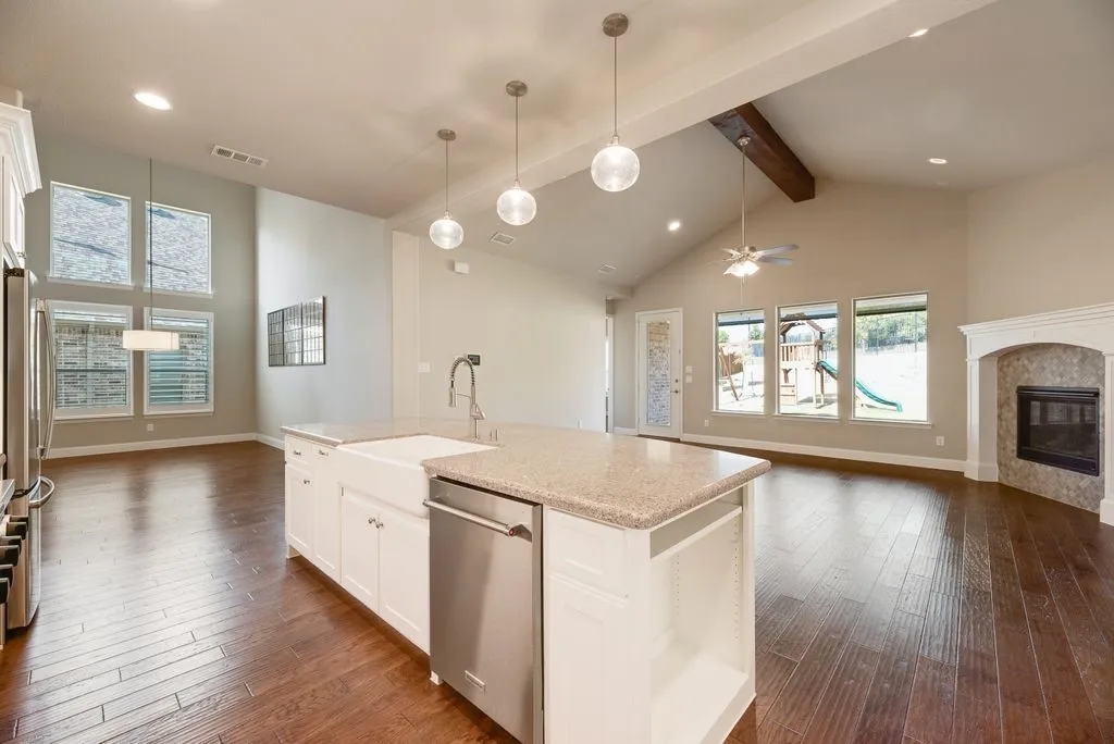 Spacious living area featuring wood-finish flooring, a kitchen island with an undermount farmhouse sink, vaulted ceilings with exposed beam, a fireplace with a decorative mantel, and multiple large windows