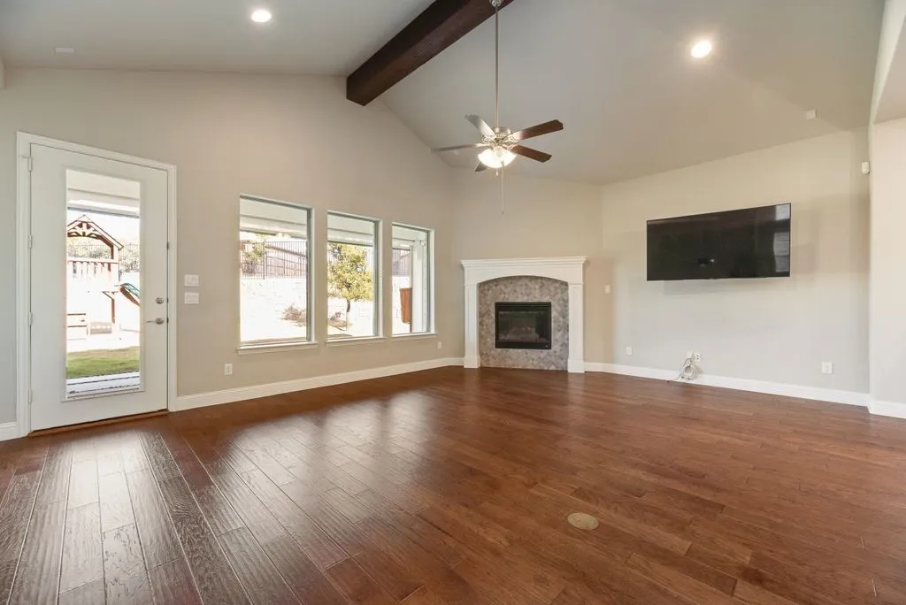 Vaulted ceiling with wood beam, wood-finish flooring, fireplace with stone surround, ceiling fan, and multiple windows