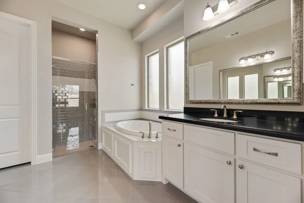 White cabinetry vanity with dark countertop, large framed mirror, corner soaking tub, walk-in shower with subway tile, and tile flooring