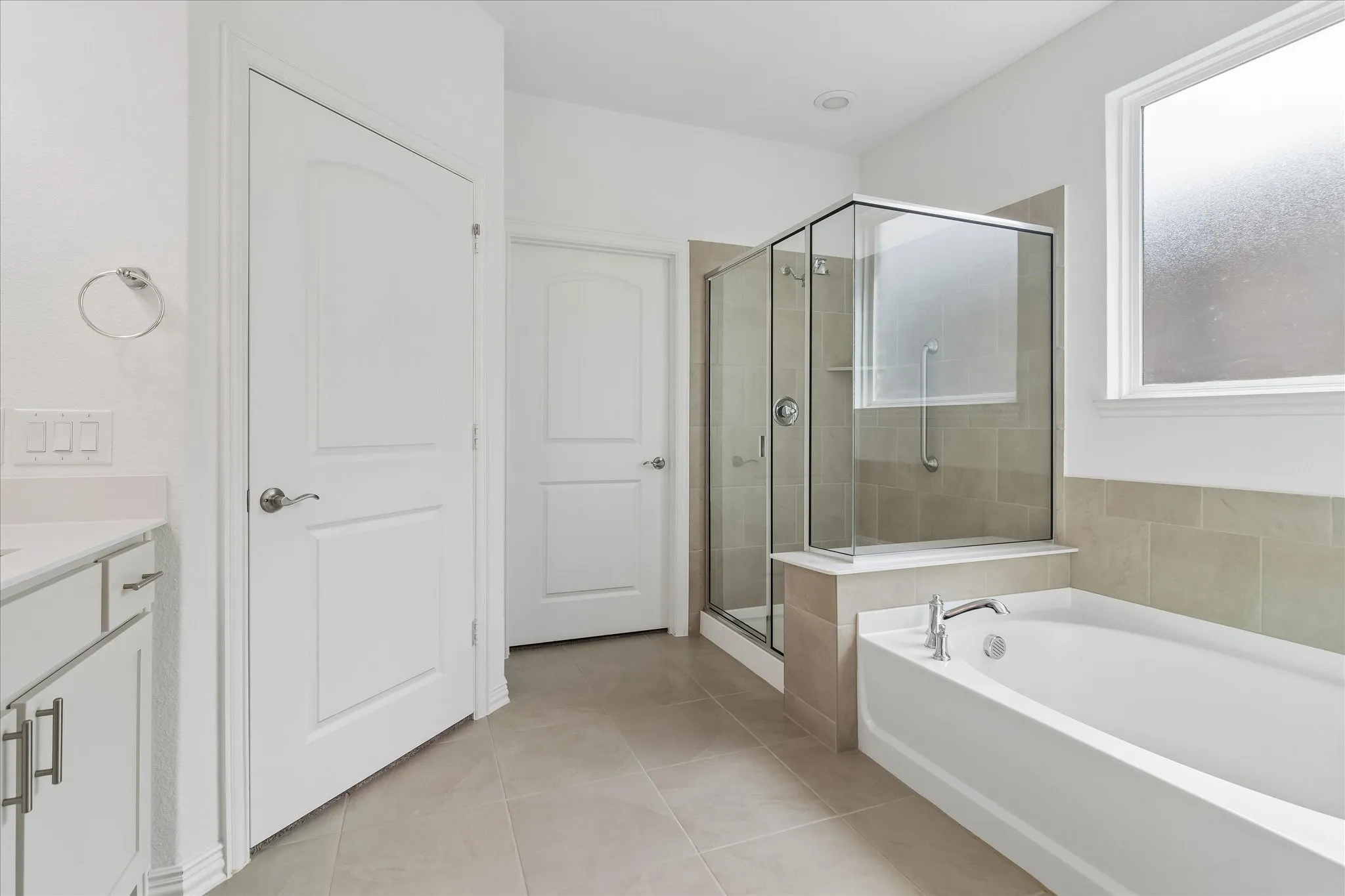 White soaking tub with chrome fixtures, glass-enclosed shower, frosted window, and white cabinetry