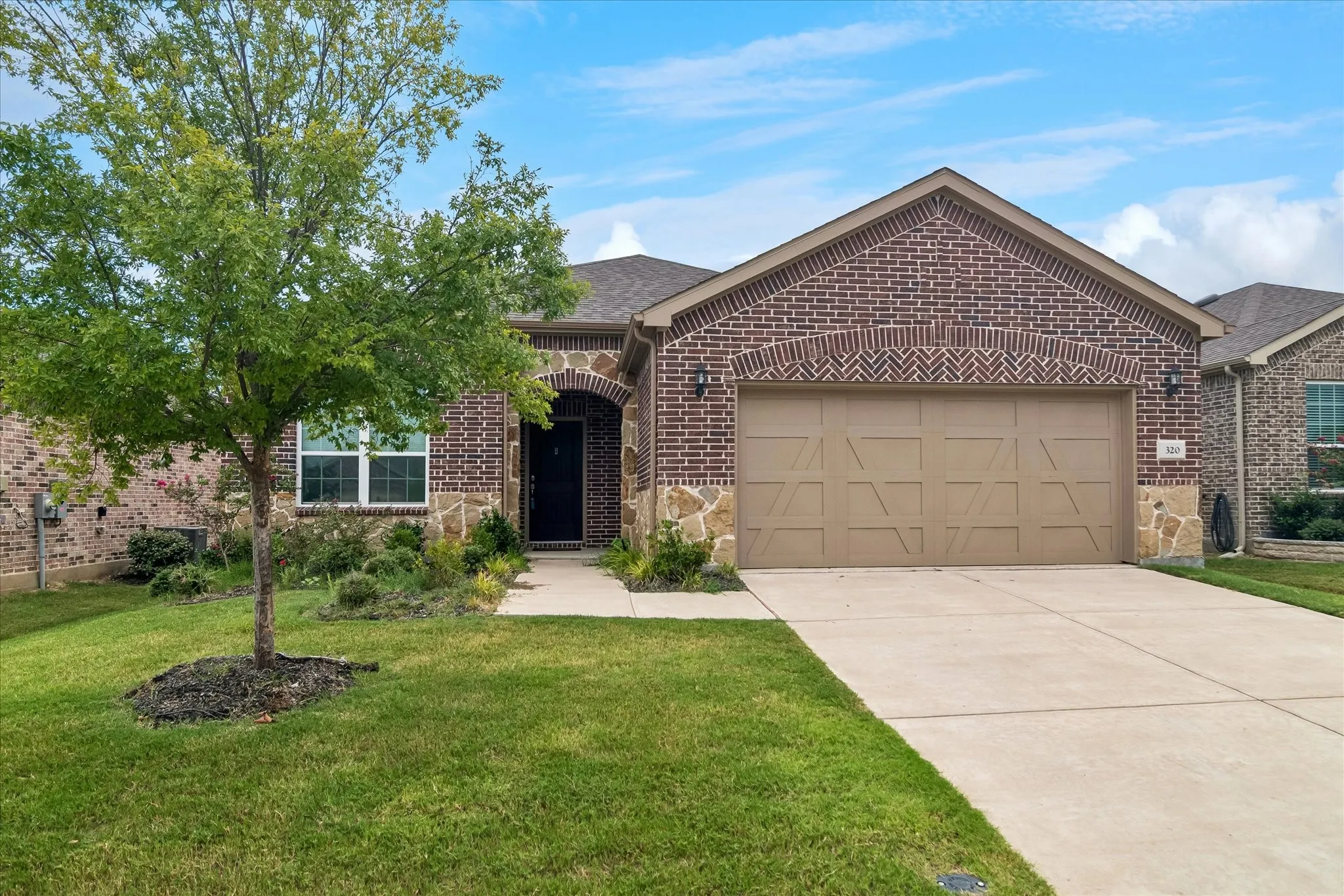 Brick and stone exterior featuring an arched entryway, attached two-car garage, concrete driveway, and a front yard with mature landscaping