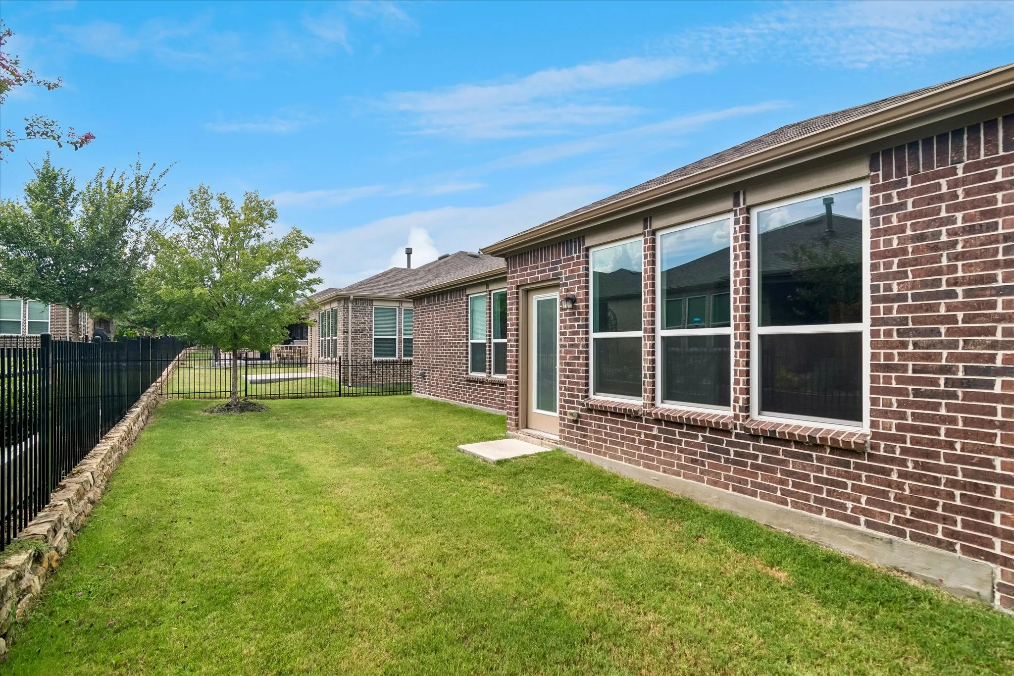 Expansive green lawn with mature trees and a black metal fence, complemented by a brick exterior featuring multiple windows and a glass-paneled door
