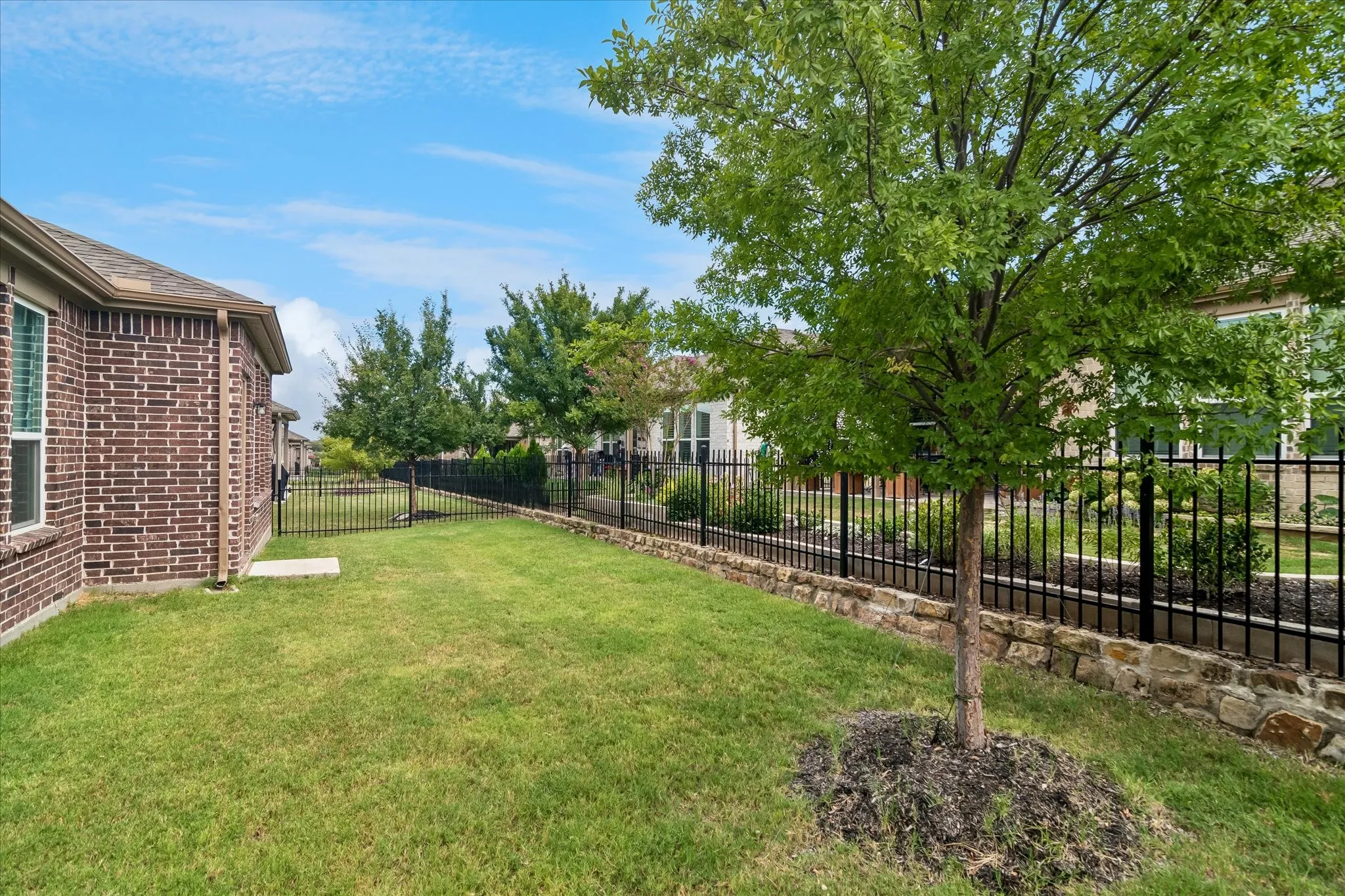 Manicured lawn, brick exterior, black metal fencing, stone retaining wall, and mature trees