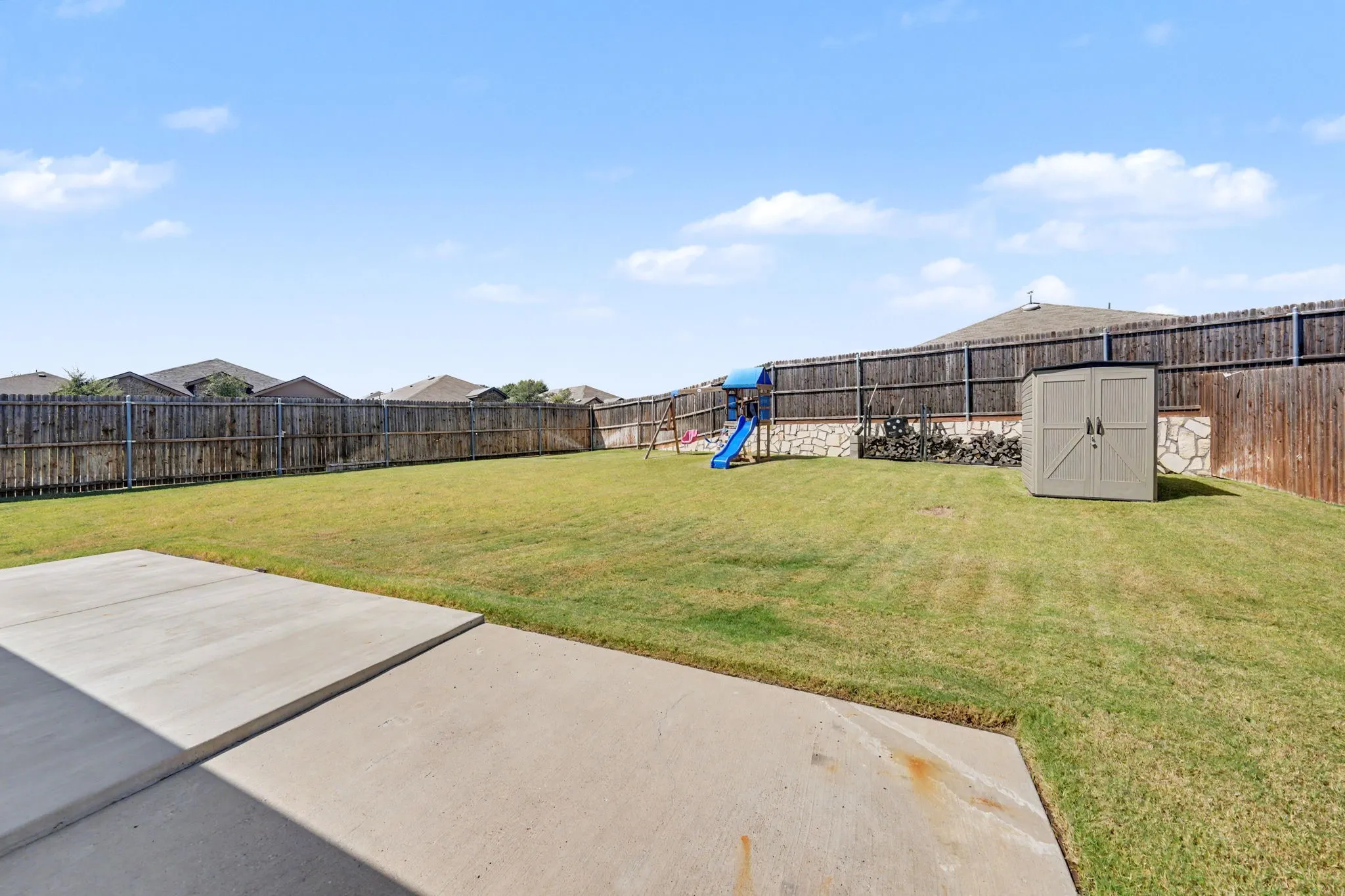 Expansive backyard featuring a concrete patio, a privacy fence, a storage shed, and a stone retaining wall