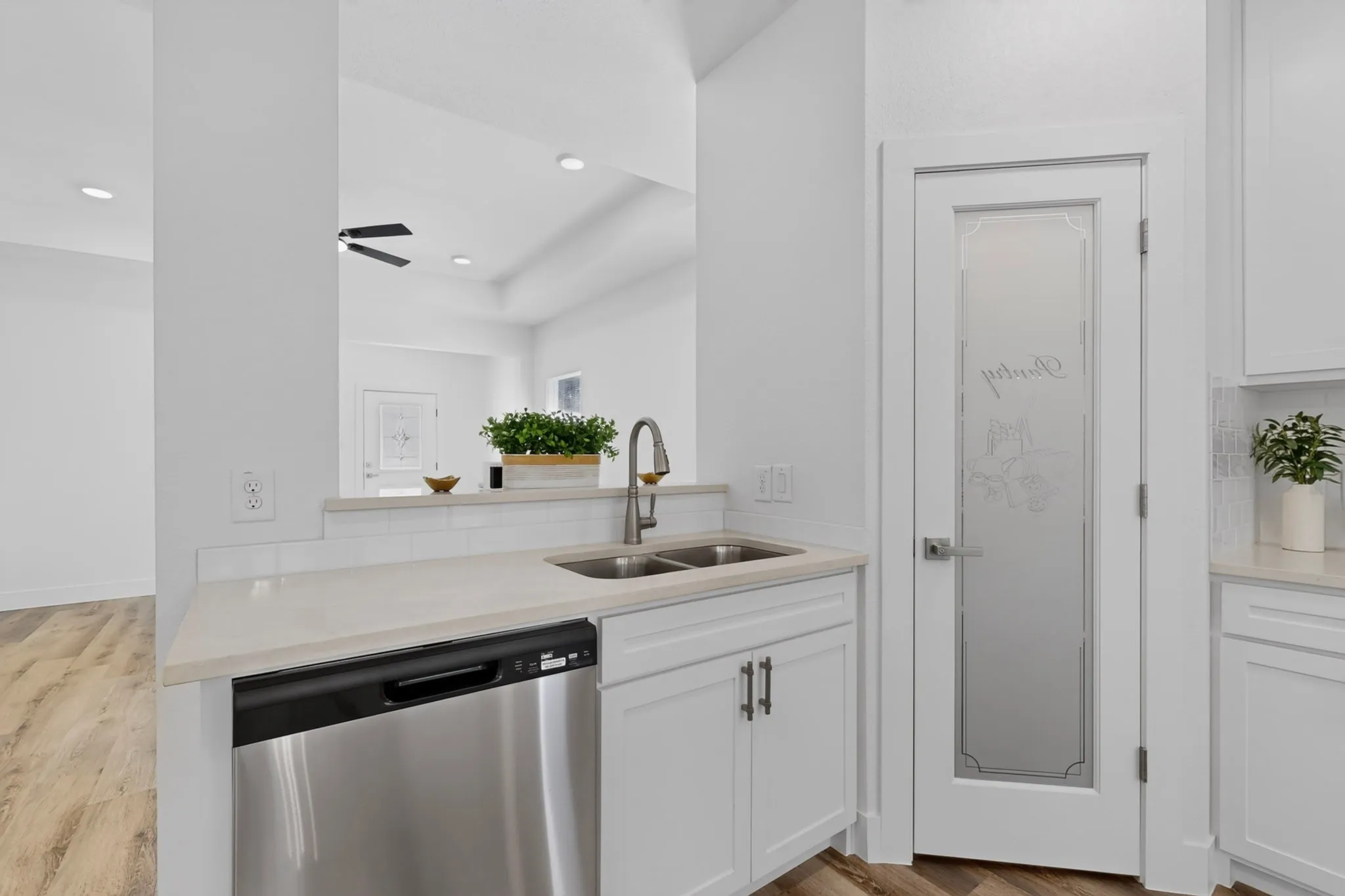 Stainless steel dishwasher, double basin sink with gooseneck faucet, lquartz countertops, white cabinetry with dark hardware, and a pantry door with frosted glass.