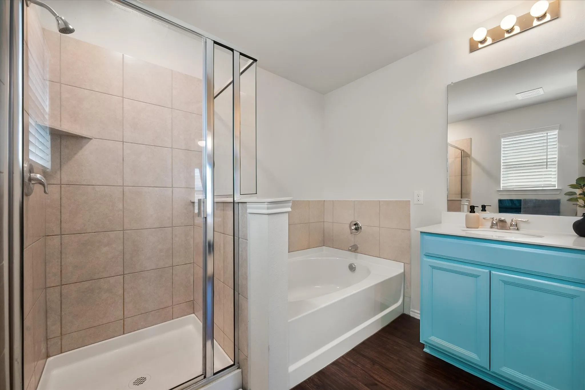 Spacious bathroom featuring a glass-enclosed shower with tile surround, a corner soaking tub with tile surround, a single vanity with a light blue cabinet, and wood-finish flooring