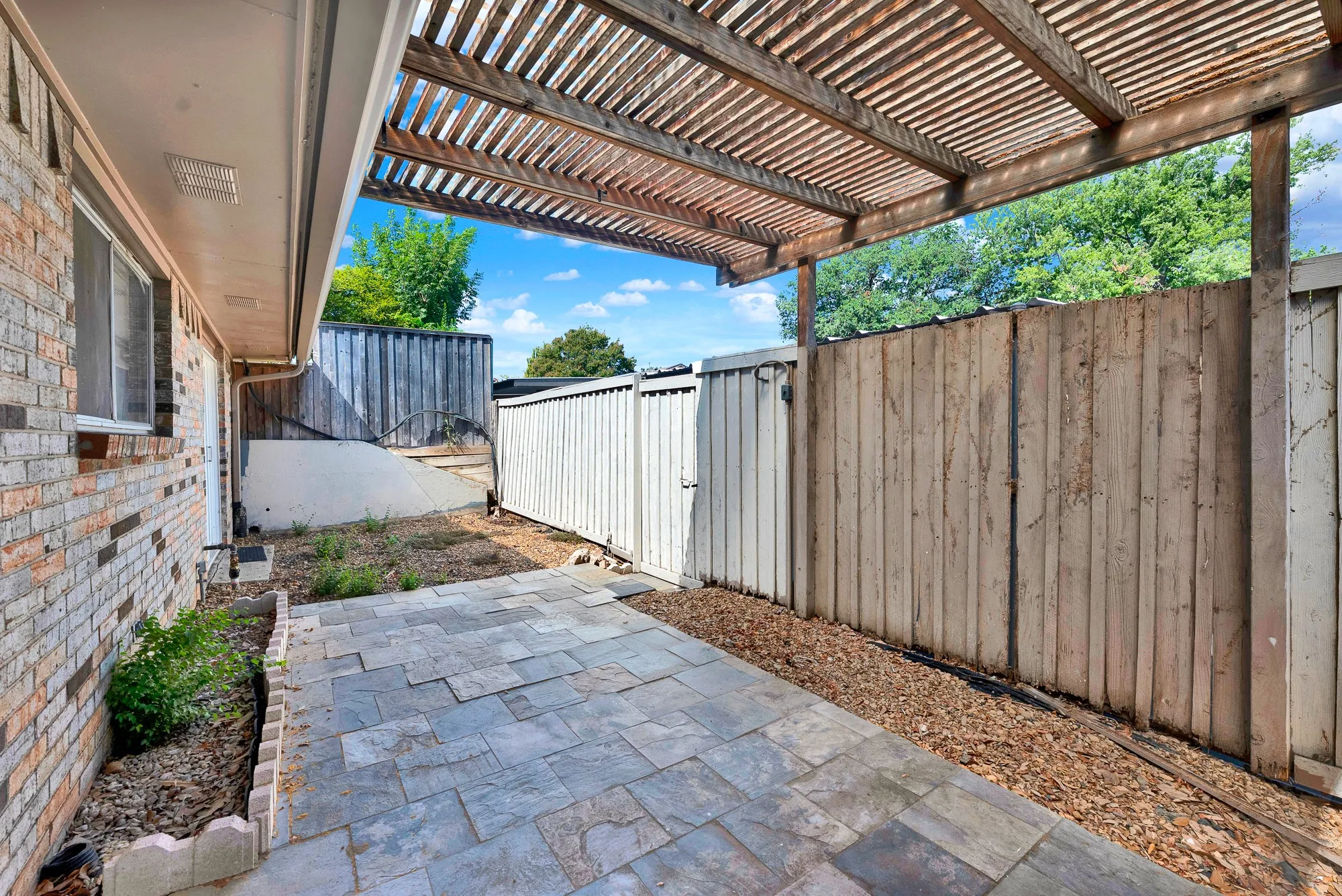 Brick exterior with a window, wood slat pergola, stone paver patio, wood fencing, and a planter area with mulch